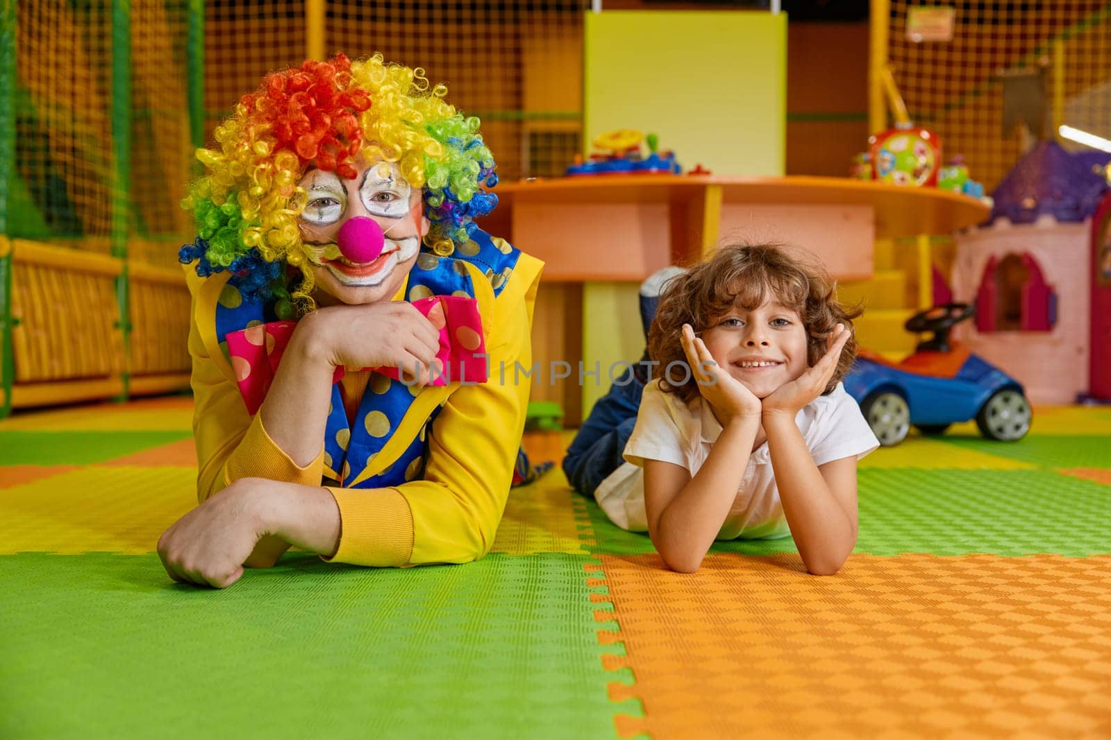 Portrait of happy smiling clown and little boy child lying on floor of indoor playground. Show entertainment, animation program for kids to celebrate birthday party with fun and joy