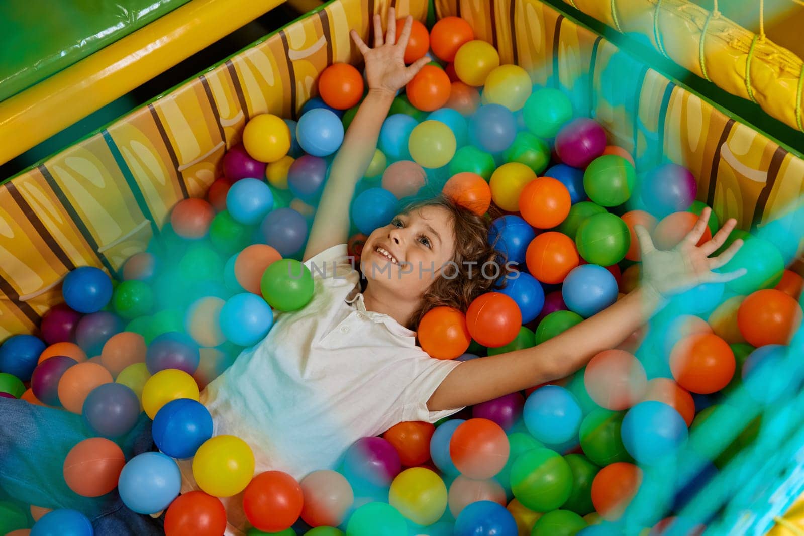 Happy boy child playing in basin filled with multi colored plastic ball. Excited cheerful kid having fun in big dry paddling pool at indoor playroom