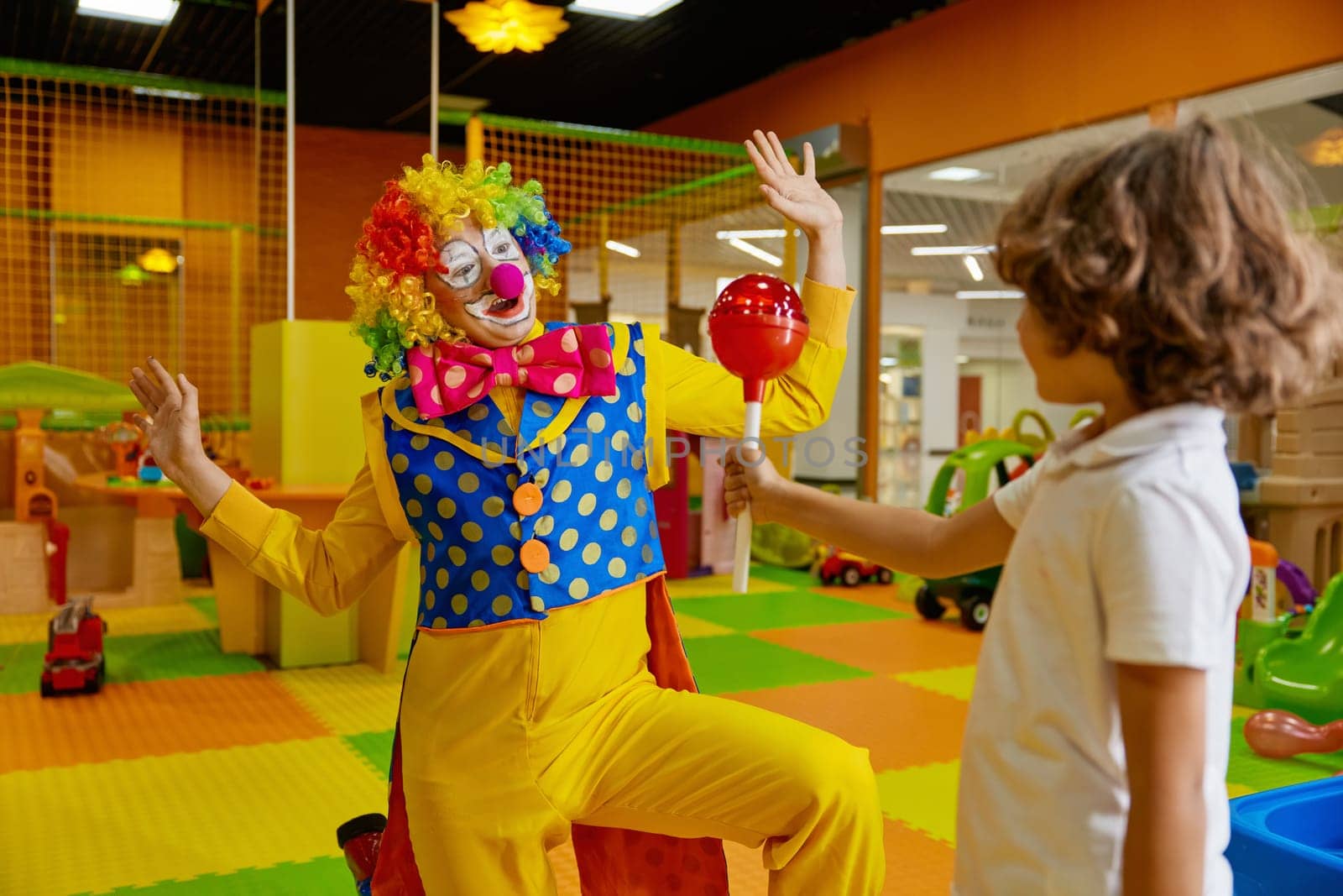 Birthday celebration at kids playroom with animation program concept. Little boy giving huge lollypop candy for surprised clown