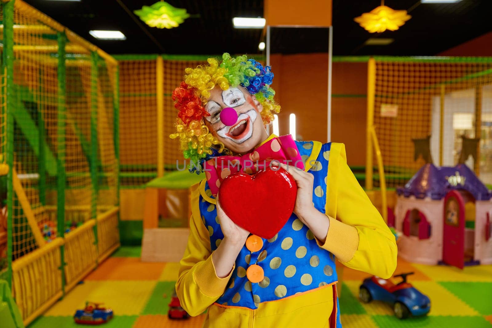 Portrait of cute funny clown artist wearing colorful wig and costume holding heart showing love and adoration feeling excitement of his professional occupation