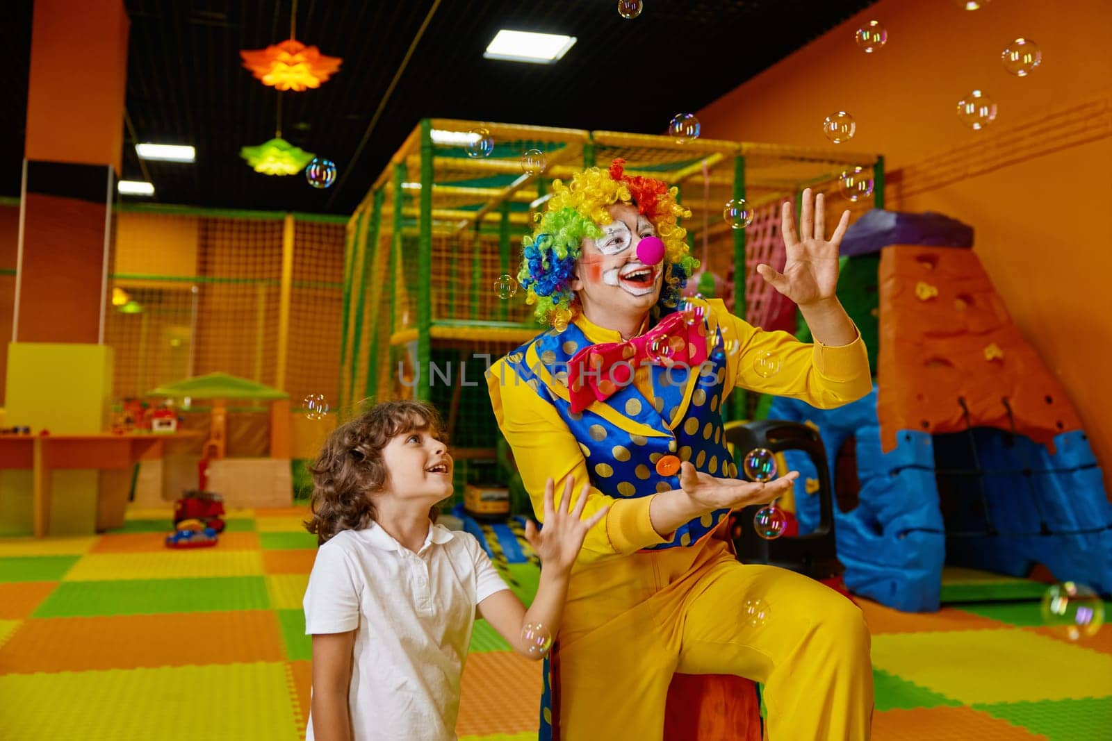 Little preschool boy and funny clown playing with flying soap bubbles. Concept of birthday entertainment at indoor city center