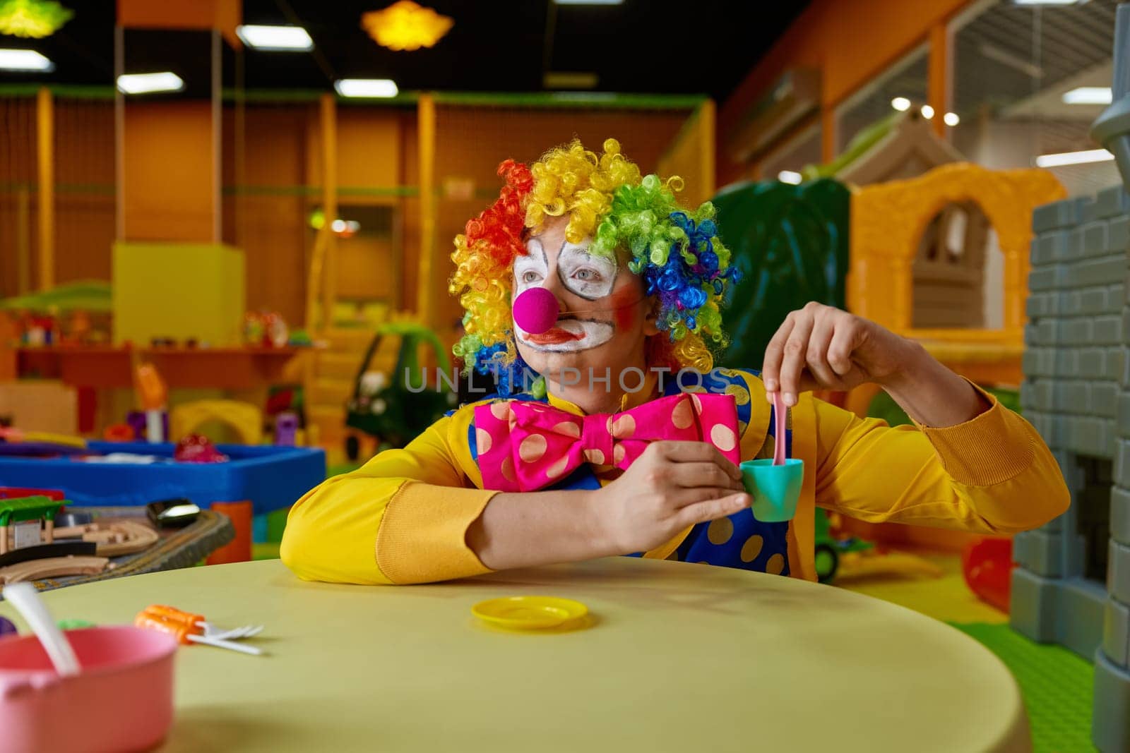 Funny clown wearing multi-colored wig and costume playing with toy tea set at kids playroom. Birthday party animation show concept