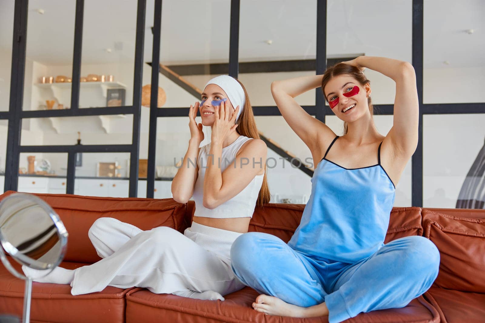 Young woman friends in pajamas applying eye patch taking care of face skin while sitting on sofa at home living room. Concept of youth, friendship party and relaxation