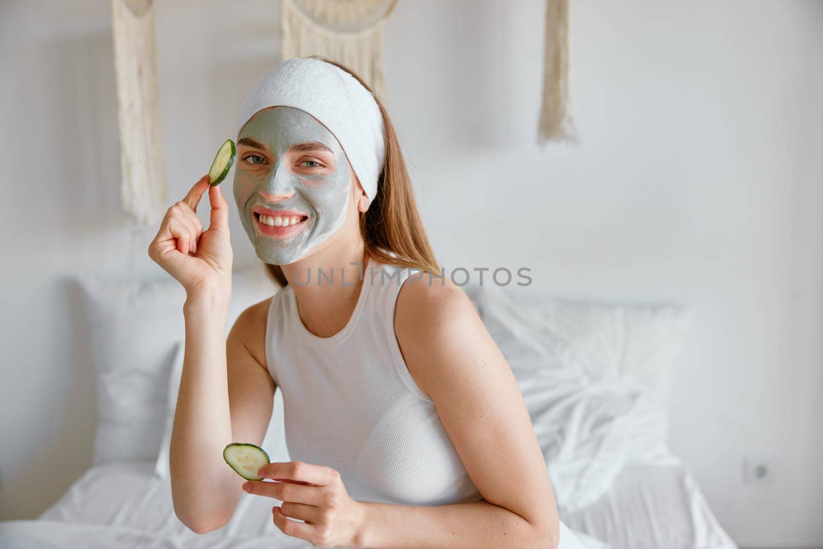 Portrait of young smiling woman enjoying home spa procedure. Millennial female applying clay mask and cucumber eye patches for skin care. Beauty treatment concept
