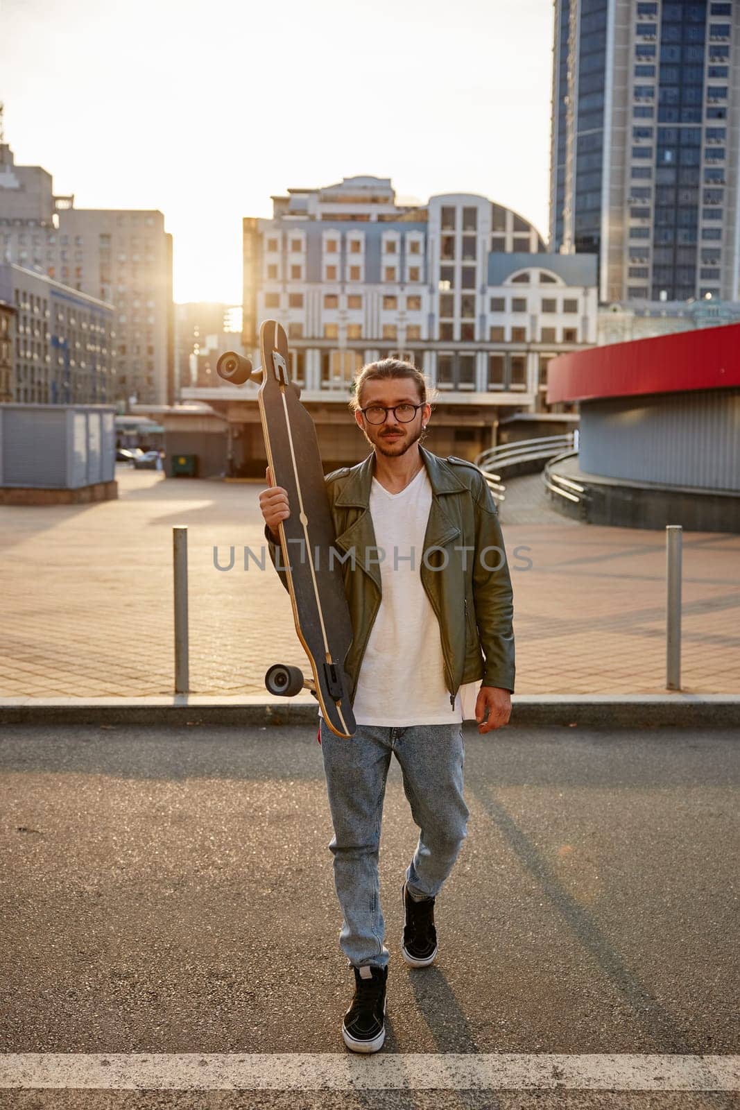 Full-length portrait of hipster guy skateboarder over urban background. Casual extreme longboard rider outside walking on camera