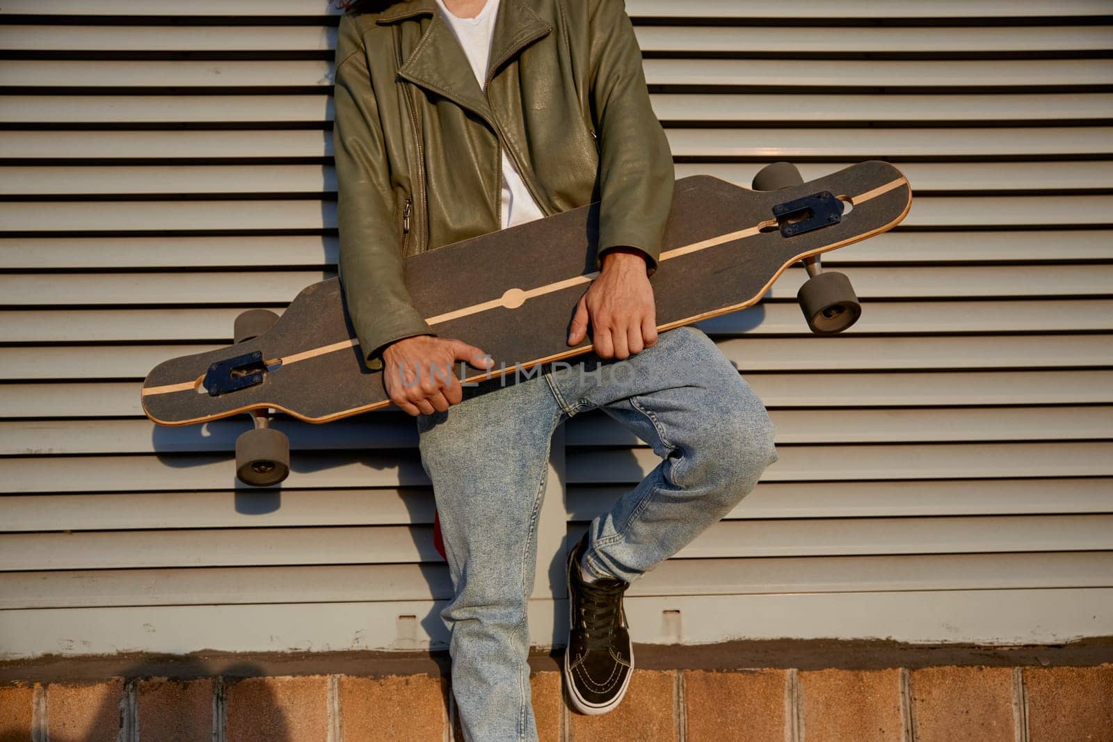 Crop of urban skater man posing for camera with longboard standing in front of street building wall