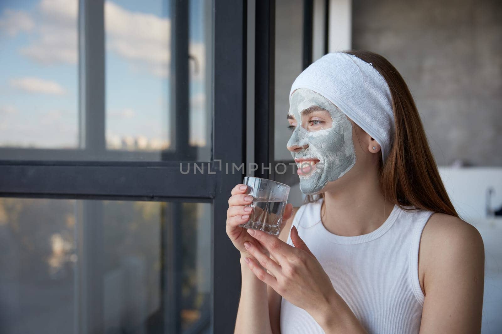 Young attractive woman morning routine for health and beauty. Charming girl with cosmetics facial mask drinking water from glass while dreamily looking in window. Closeup female portrait