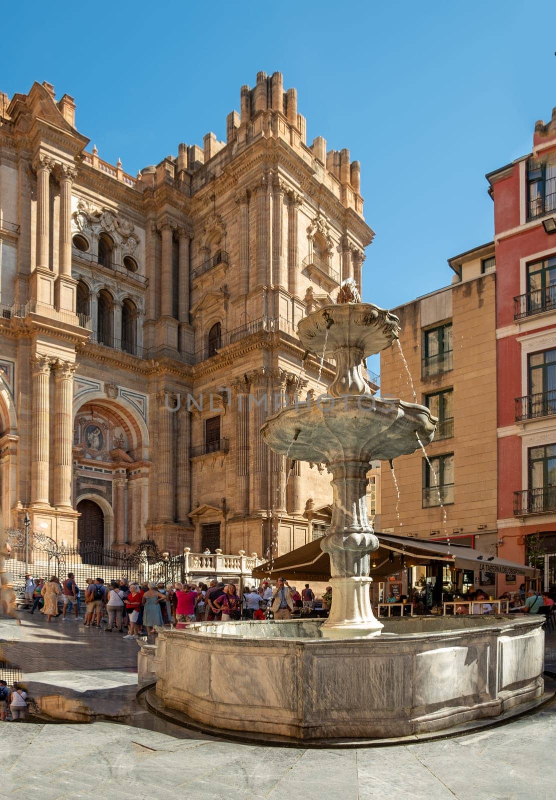 Málaga, Spain - September 28,2023: Facade of the Malaga Cathedral or Santa Iglesia Catedral Basílica de la Encarnación, with a fountain and tourists in the town square in the foreground.