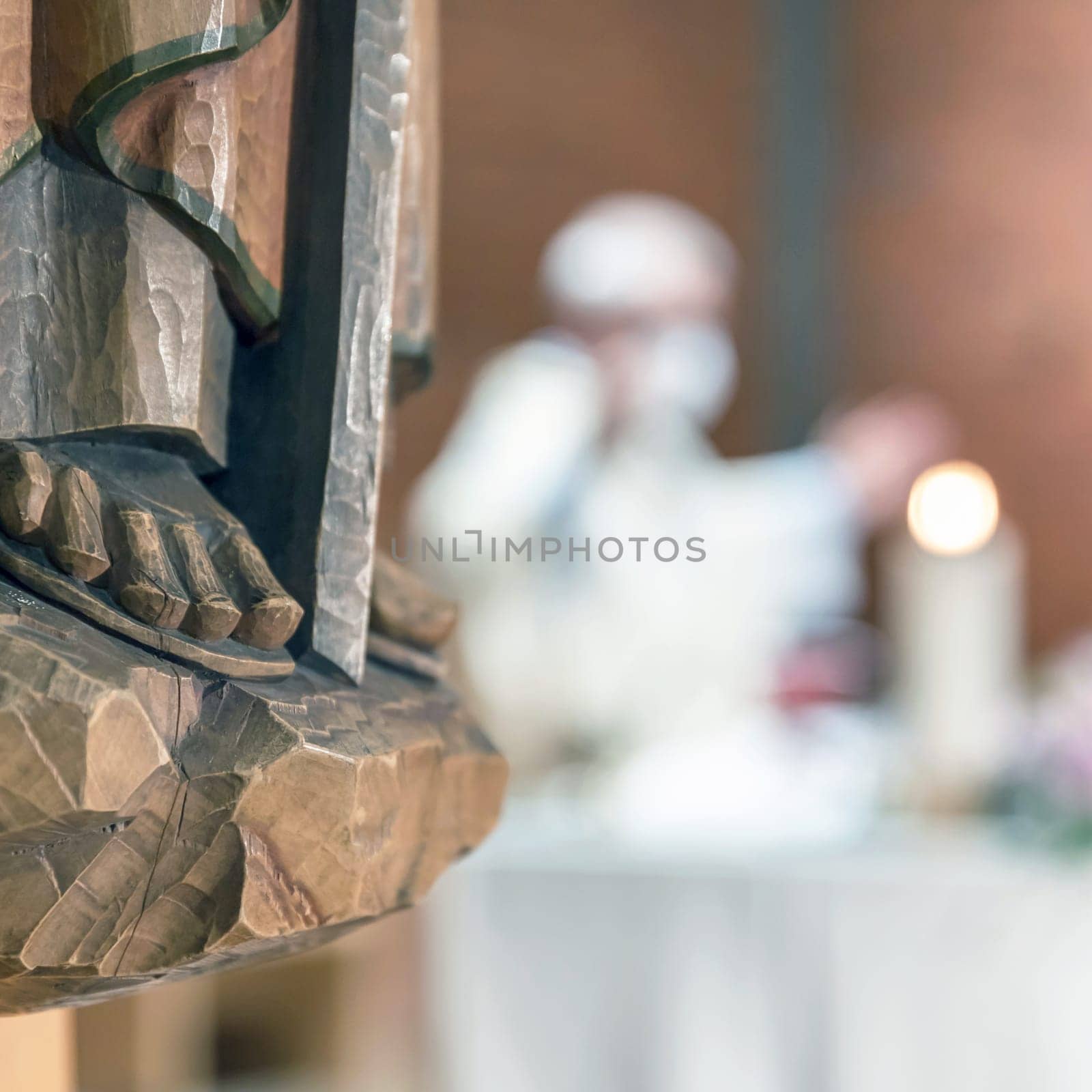 Holy wooden statue. Carved wooden feet, detail of wooden statue of a Saint. In the blurred background a priest celebrates Mass.
