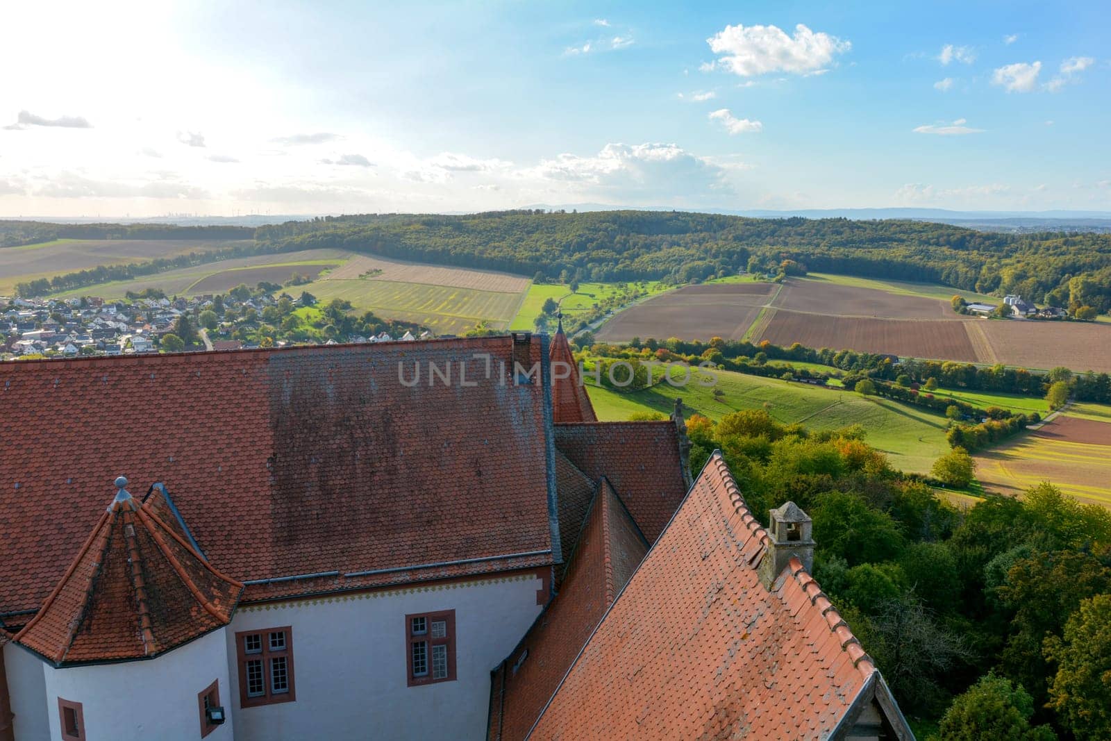 Top view of the roof of an old castle and a green landscape with houses of a village, farmland, forest and blue sky