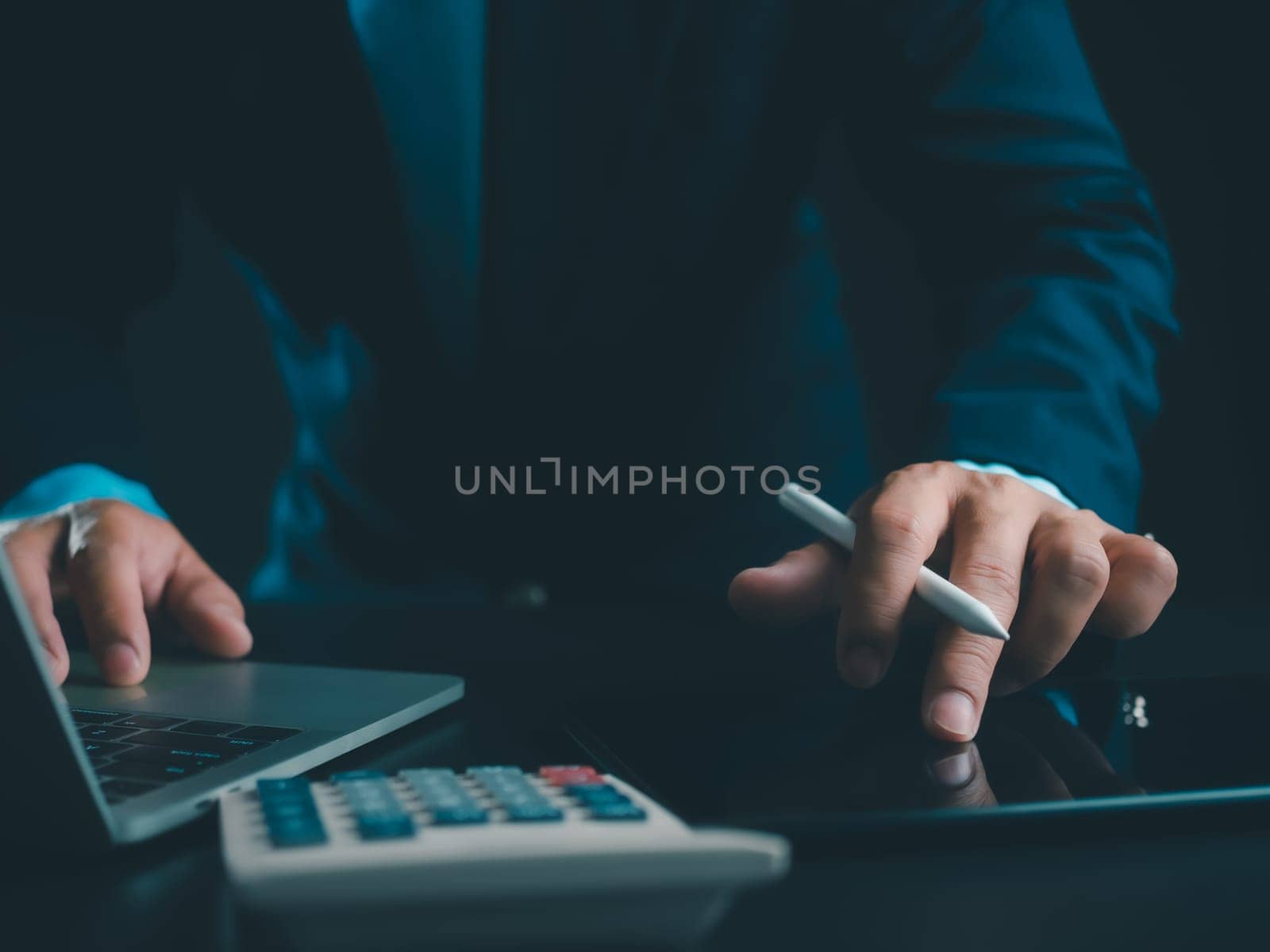 Businessman in black suit working on laptop computer and tablet and calculator, Hand Hands touch on tablet at office with dark background, Online working, Close up, Copy space.