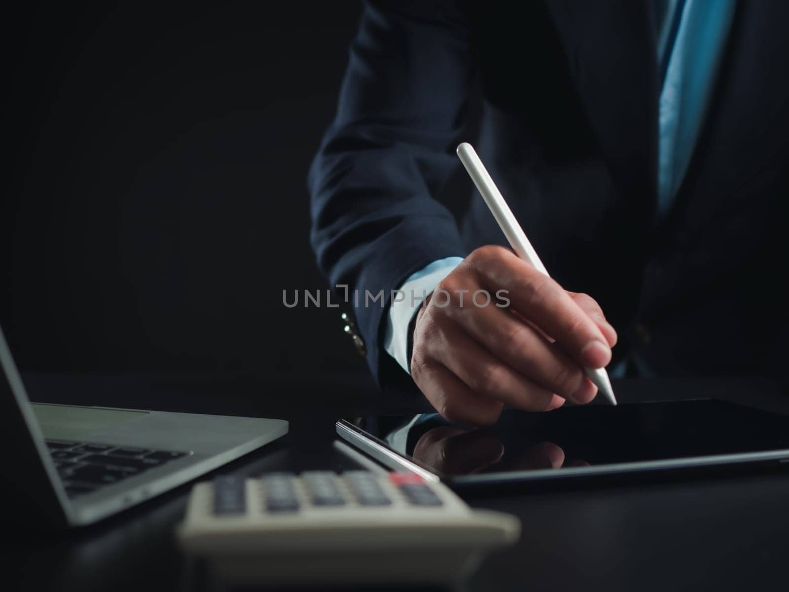Businessman in black suit working on laptop computer and tablet and calculator, Hand Hands touch on tablet at office with dark background, Online working, Close up, Copy space.