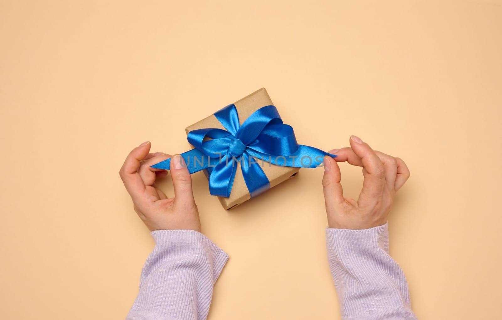 A woman's hand holds a gift box wrapped in a blue silk ribbon on a beige background, top view