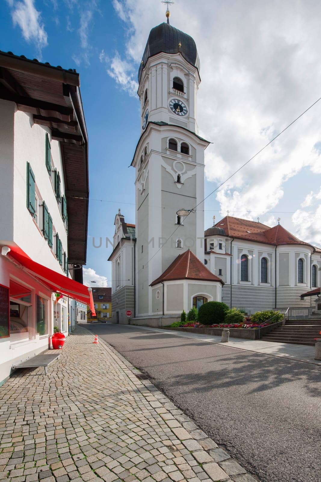 North-side wide angle view of the St. Andreas Church with bell tower in Nesselwang / Bavaria from in front of a blue sky with clouds and lens flares in the summer of 2023.