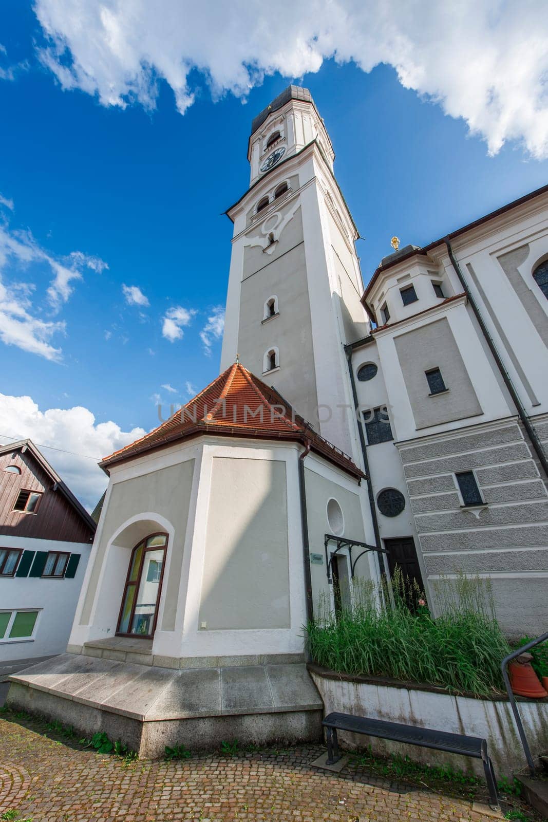 Wide-angle view of the Saint Andreas Church with bell tower and tower chapel in Nesselwang Bavaria from the northwest against a blue sky with clouds in summer 2023.