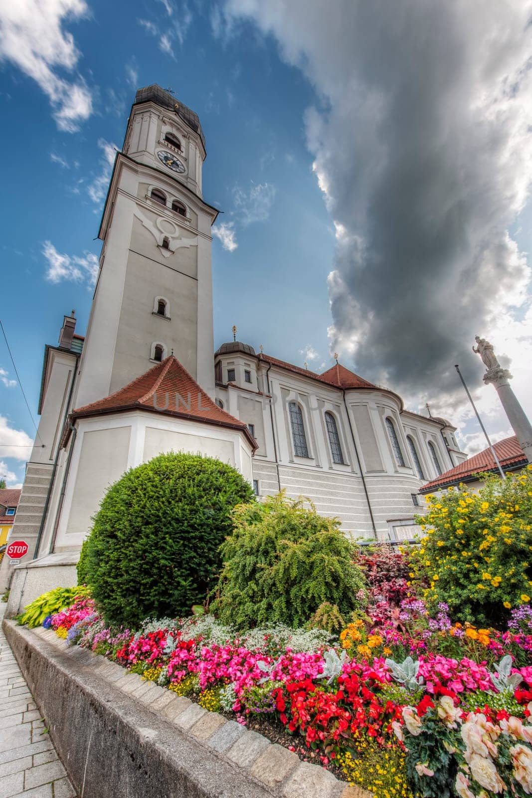 North-side extreme wide-angle view of the Saint Andreas Church with the bell tower and flower beds in front of it in Nesselwang/Bavaria against a blue sky with clouds in summer 2023.