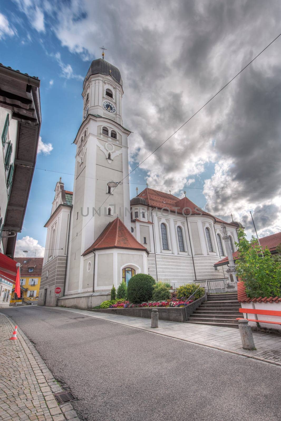North-side wide angle view of the St. Andreas Church with bell tower in Nesselwang / Bavaria from in front of a blue sky with clouds and lens flares in the summer of 2023.