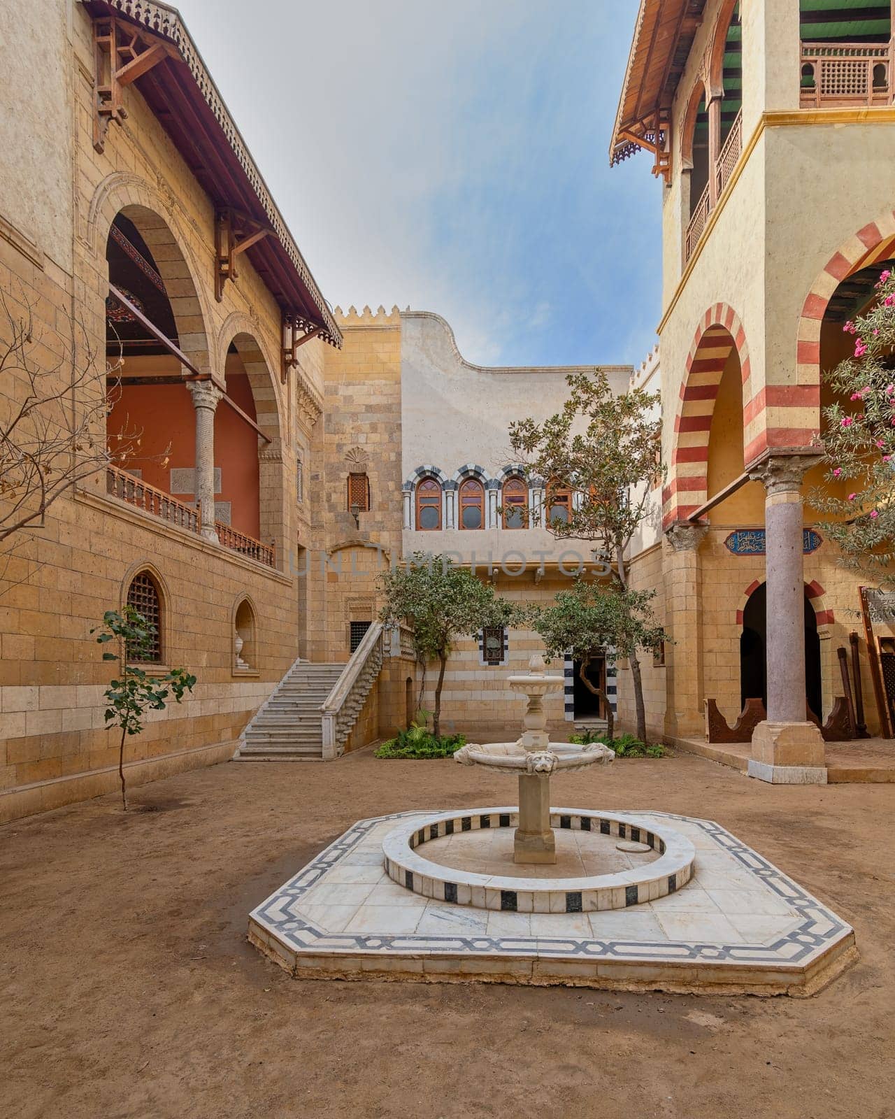 Serene courtyard showcases the timeless beauty of Mamluk architecture with high big arches and stonework with welcoming staircase leading to ornate balcony. A classic marble fountain sits at center