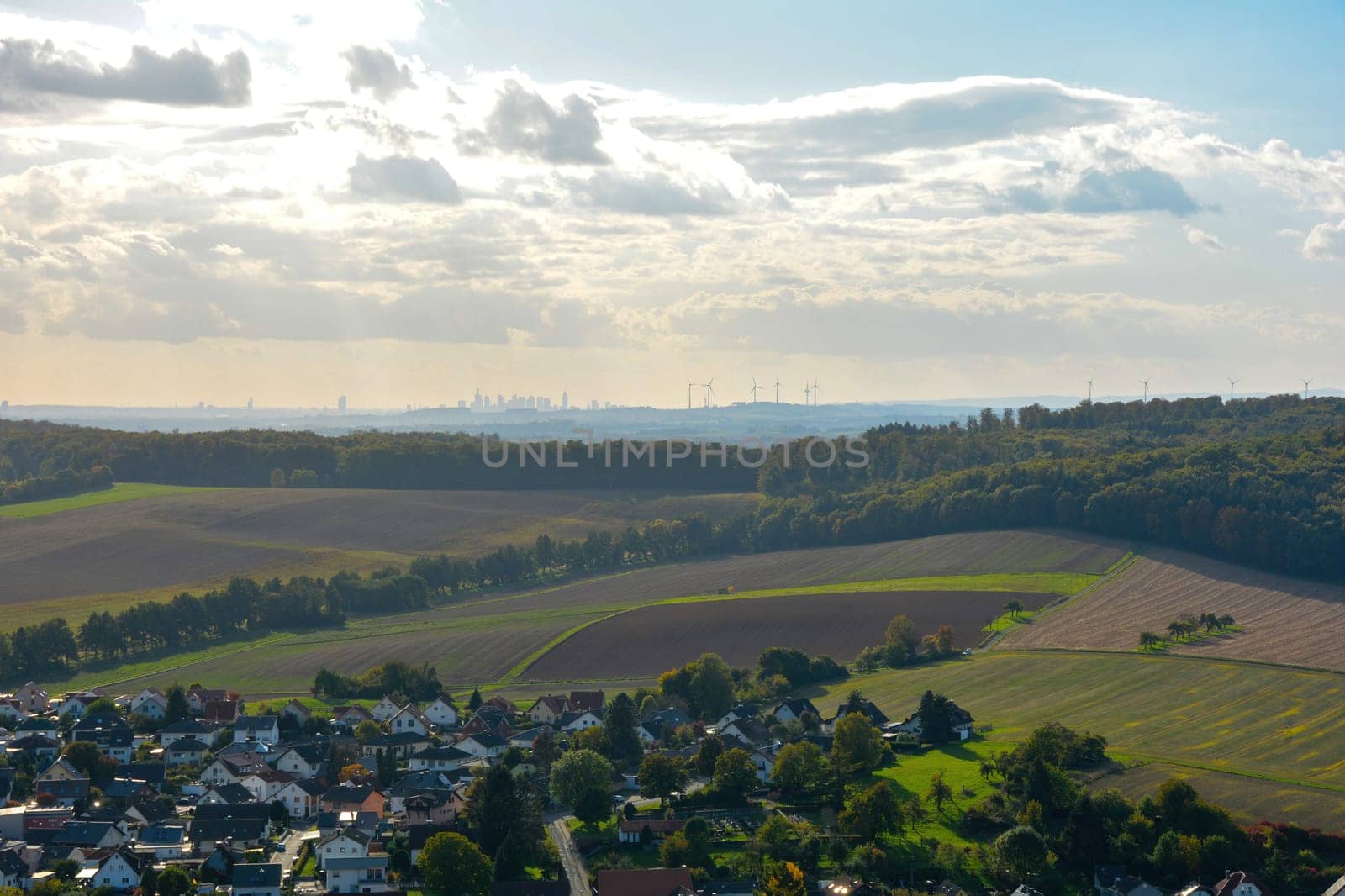Top view of a green landscape with houses of a village, farmland, forest and blue sky. In the background the skyline of the city of Frankfurt am Main, Hesse, Germany