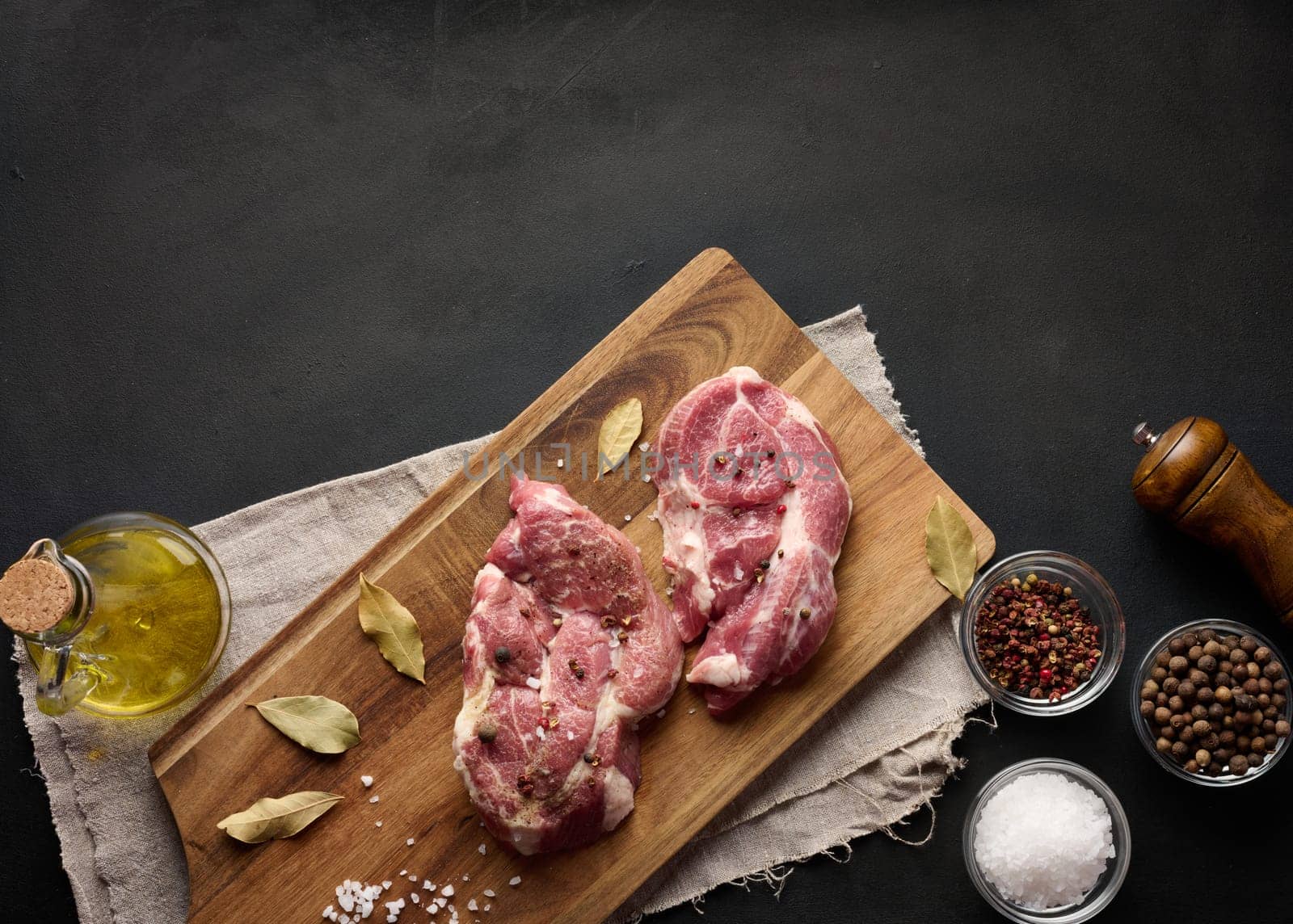 Two raw pork neck steaks on a board and spices for cooking. Top view of black table. Copy space