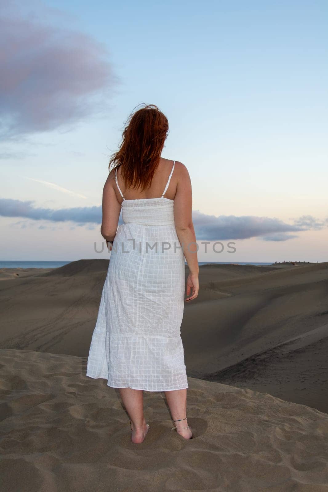 A young girl stands in a white dress in the sand dunes with sky and clouds in the late afternoon