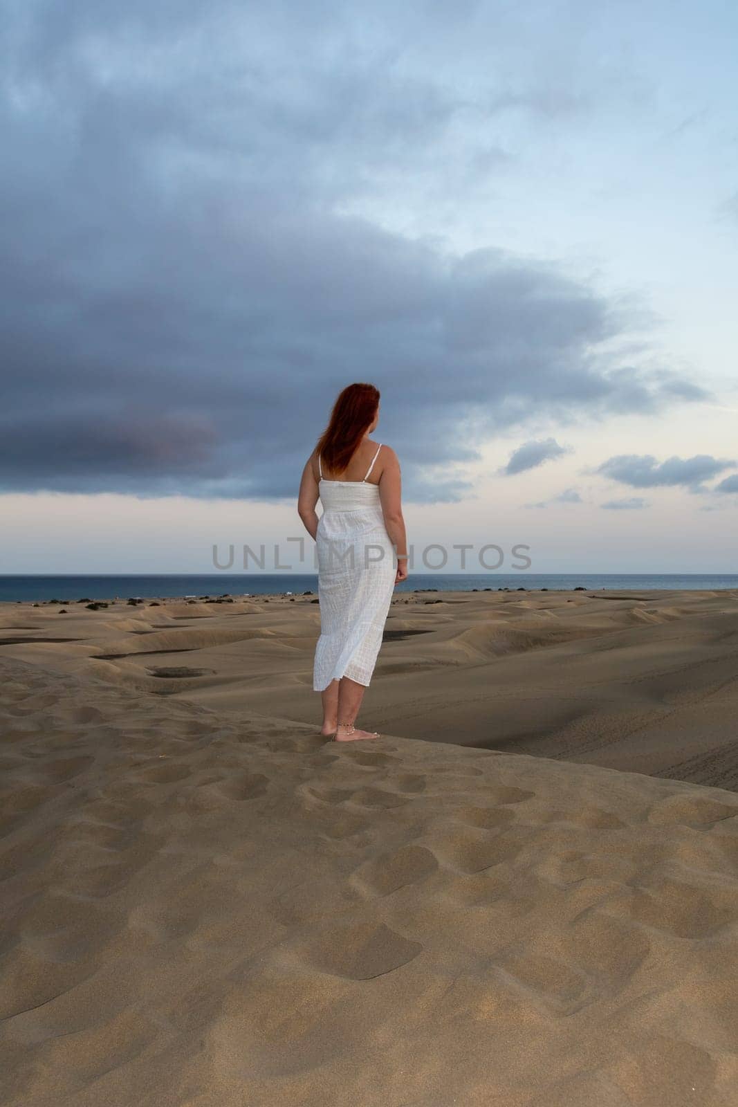A young girl stands in a white dress in the sand dunes with sky and clouds in the late afternoon