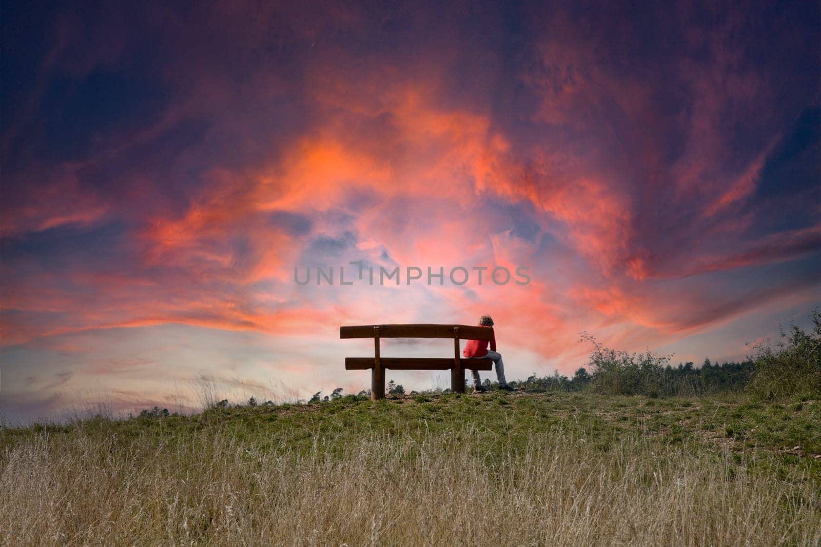 A teenager from behind sits alone on a bench in nature with lots of sky