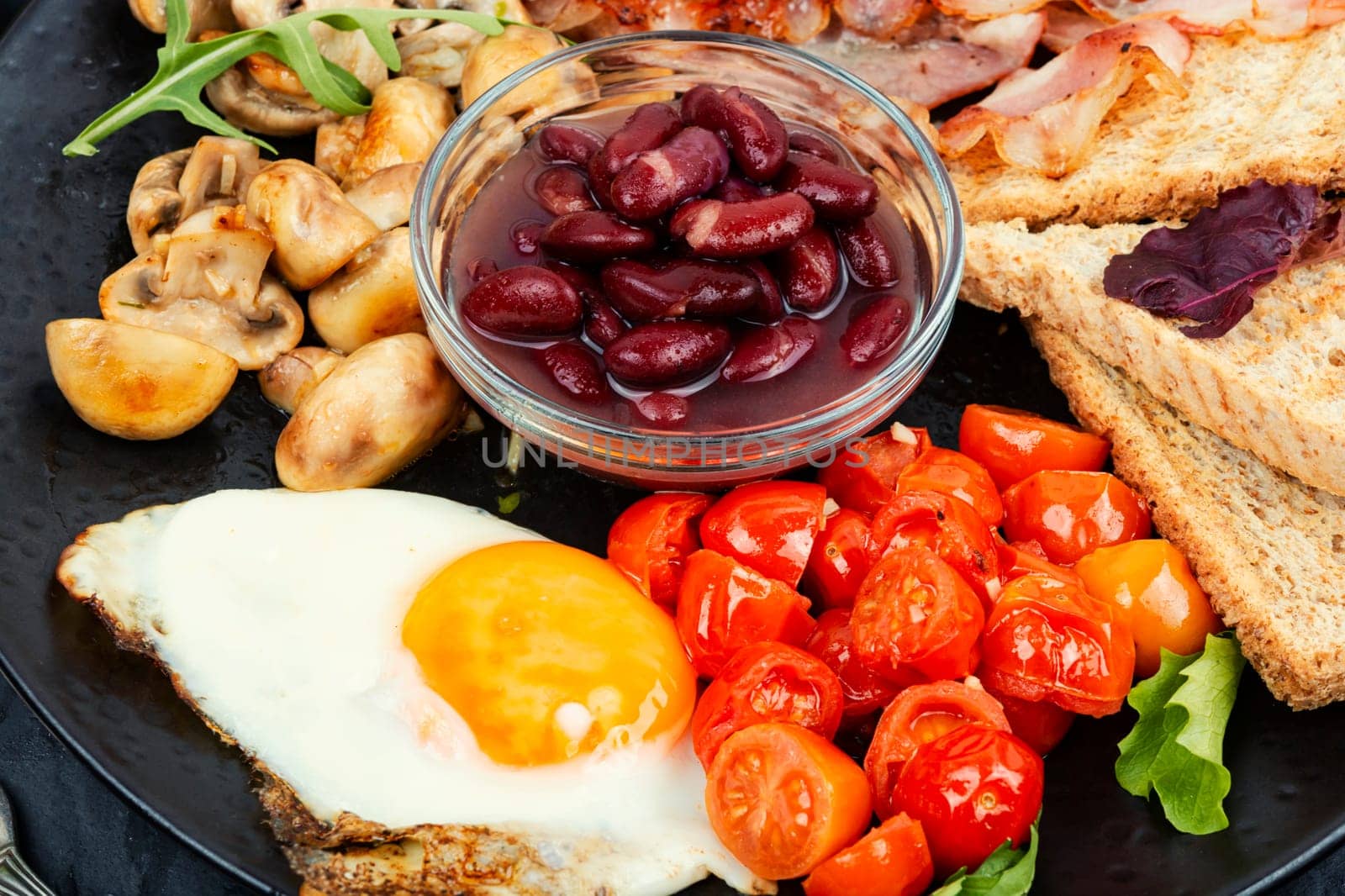 Full English breakfast of scrambled eggs, roasted bacon, toast and beans. Close up.