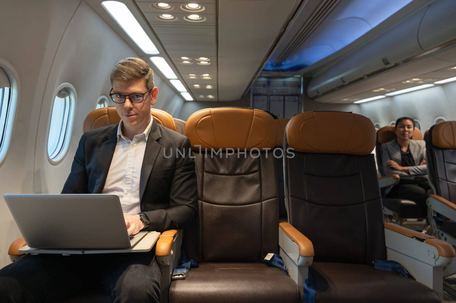 Handsome businessman with eyeglasses working on laptop computer while sitting in airplane cabin. Internet connection on board service and business travel concept.
