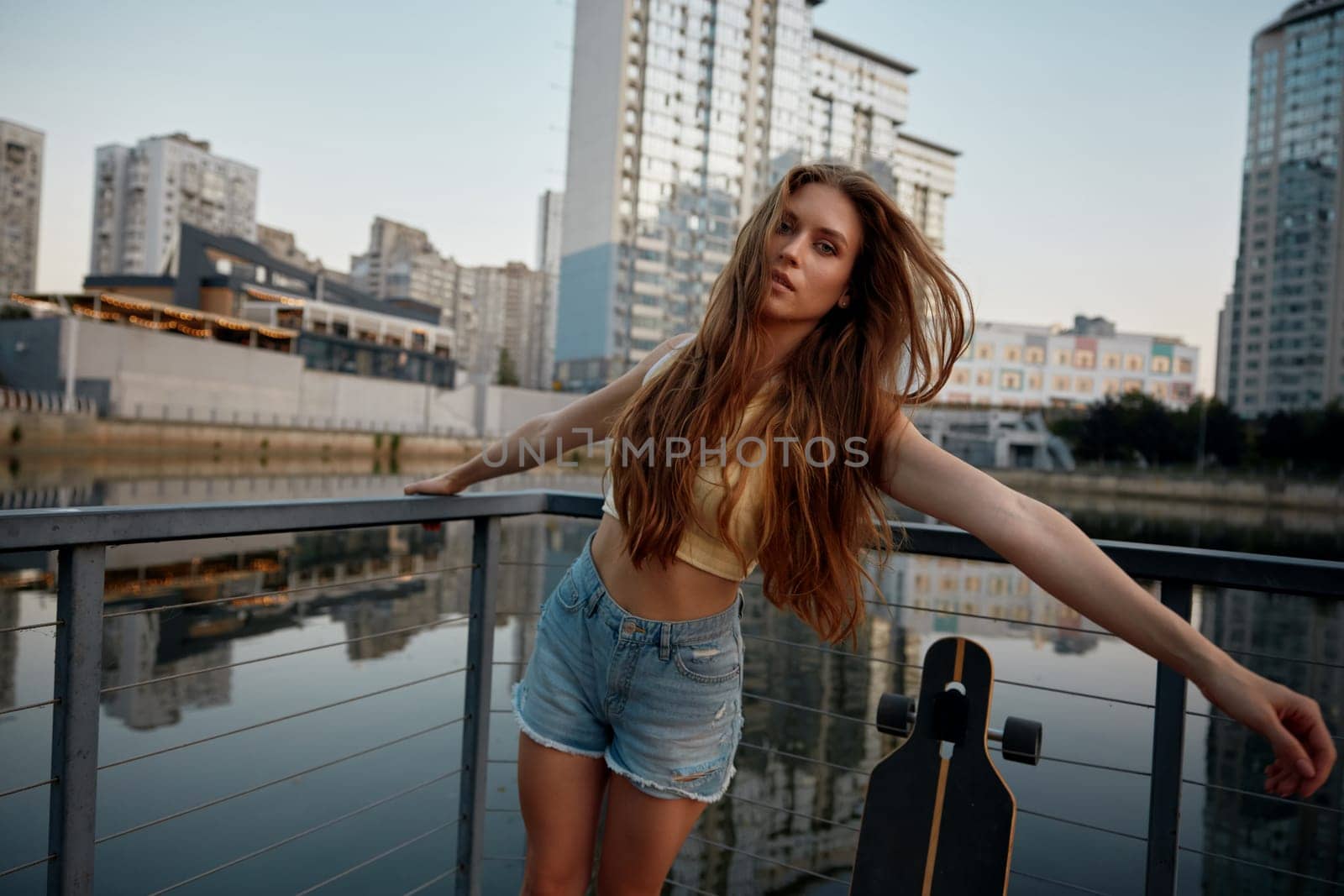 Young teenage woman enjoying time for skateboarding through city street. Beautiful female posing for camera standing with longboard over urban river in city park