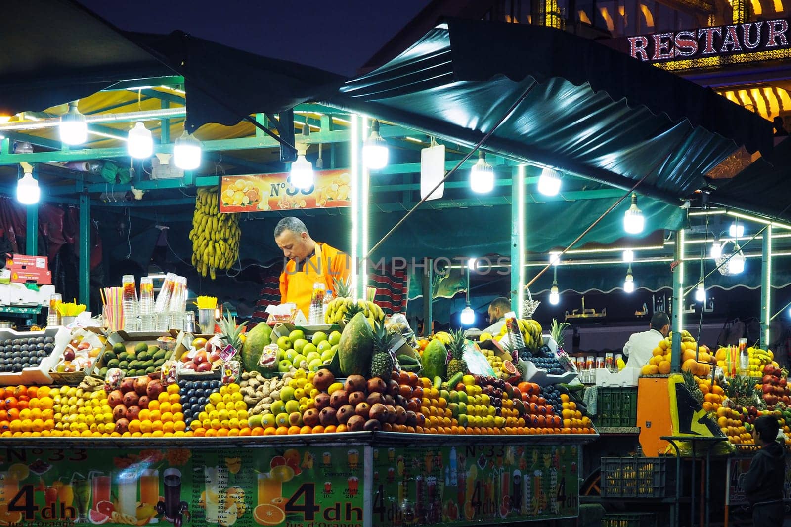 Marrakech, Morocco - January 1st, 2020: Unknown local man selling fresh fruit juice at street market stall bar in evening - heaps of fruits around
