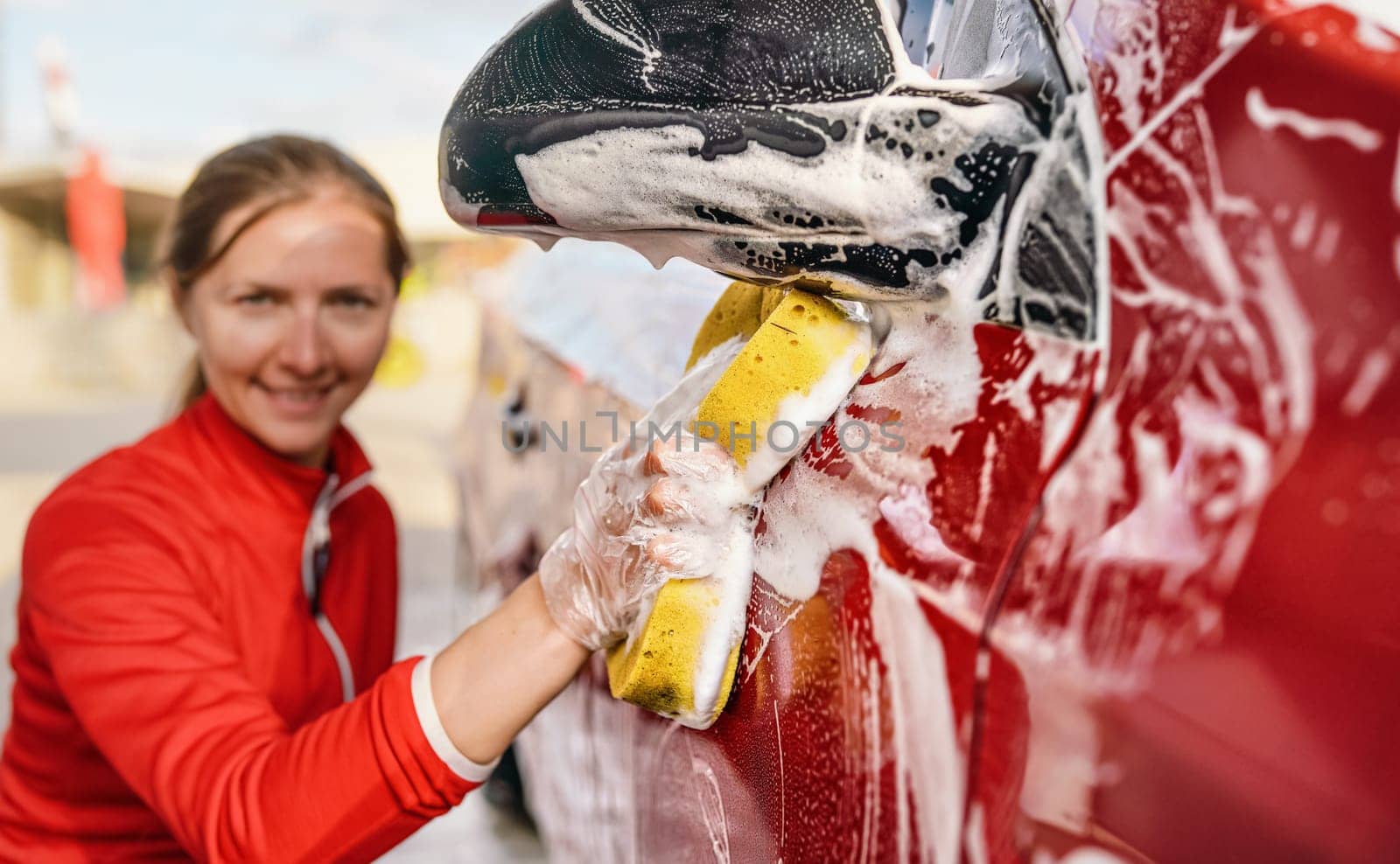 Young woman cleaning side of her car, detail on hand in glove holding yellow sponge