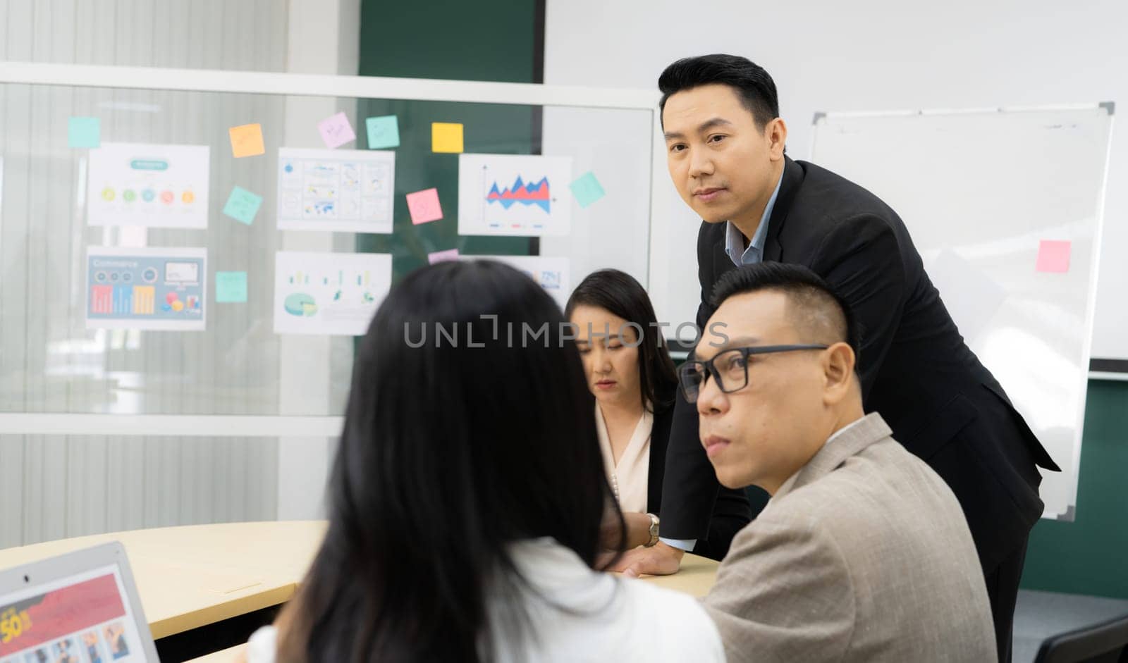 A group of people are gathered around a table with a whiteboard behind them. They are discussing something important, and one of the men is wearing glasses