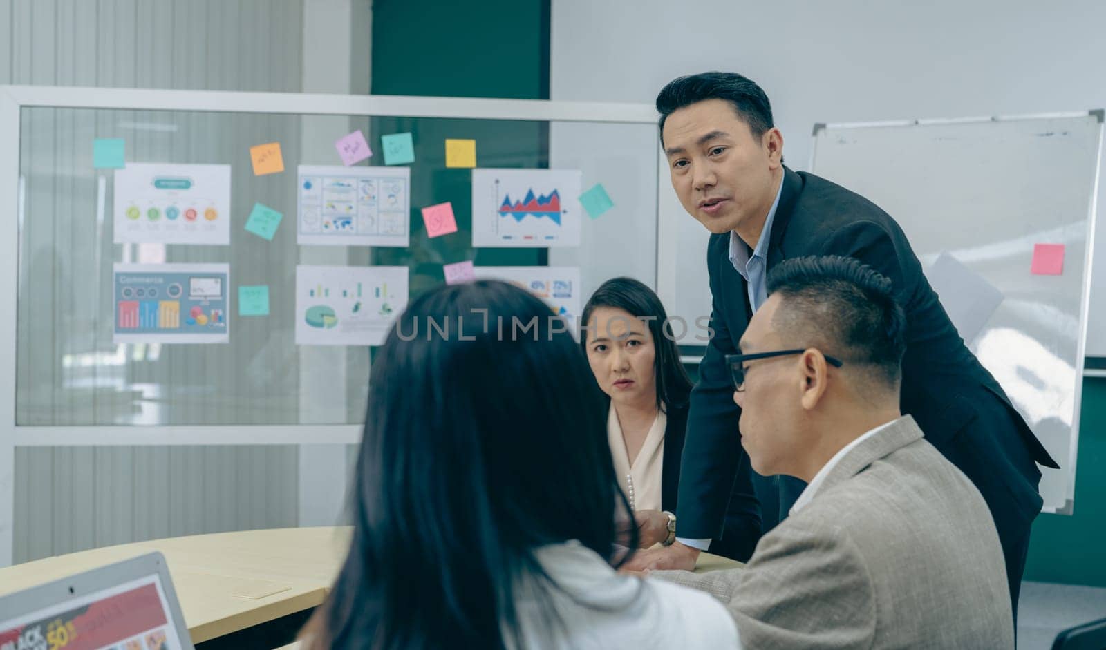 A group of people are gathered around a table with a whiteboard behind them. They are discussing something important, and there are several posters on the wall behind them