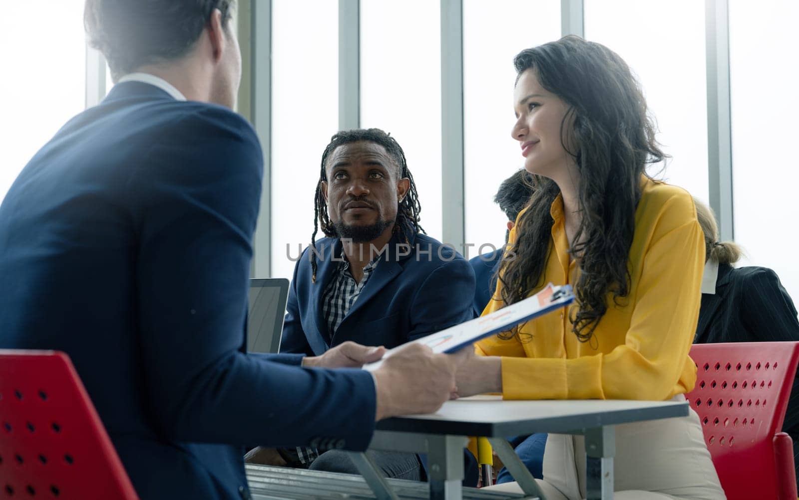 A group of people are sitting around a table, one of whom is holding a clipboard. The atmosphere seems to be professional and serious, as the group is discussing important matters