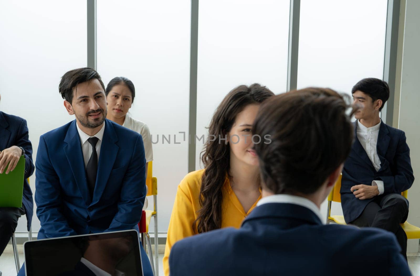 A group of people are sitting in a room, some of them wearing suits. A man in a suit is talking to a woman in a yellow dress. Scene is professional and formal