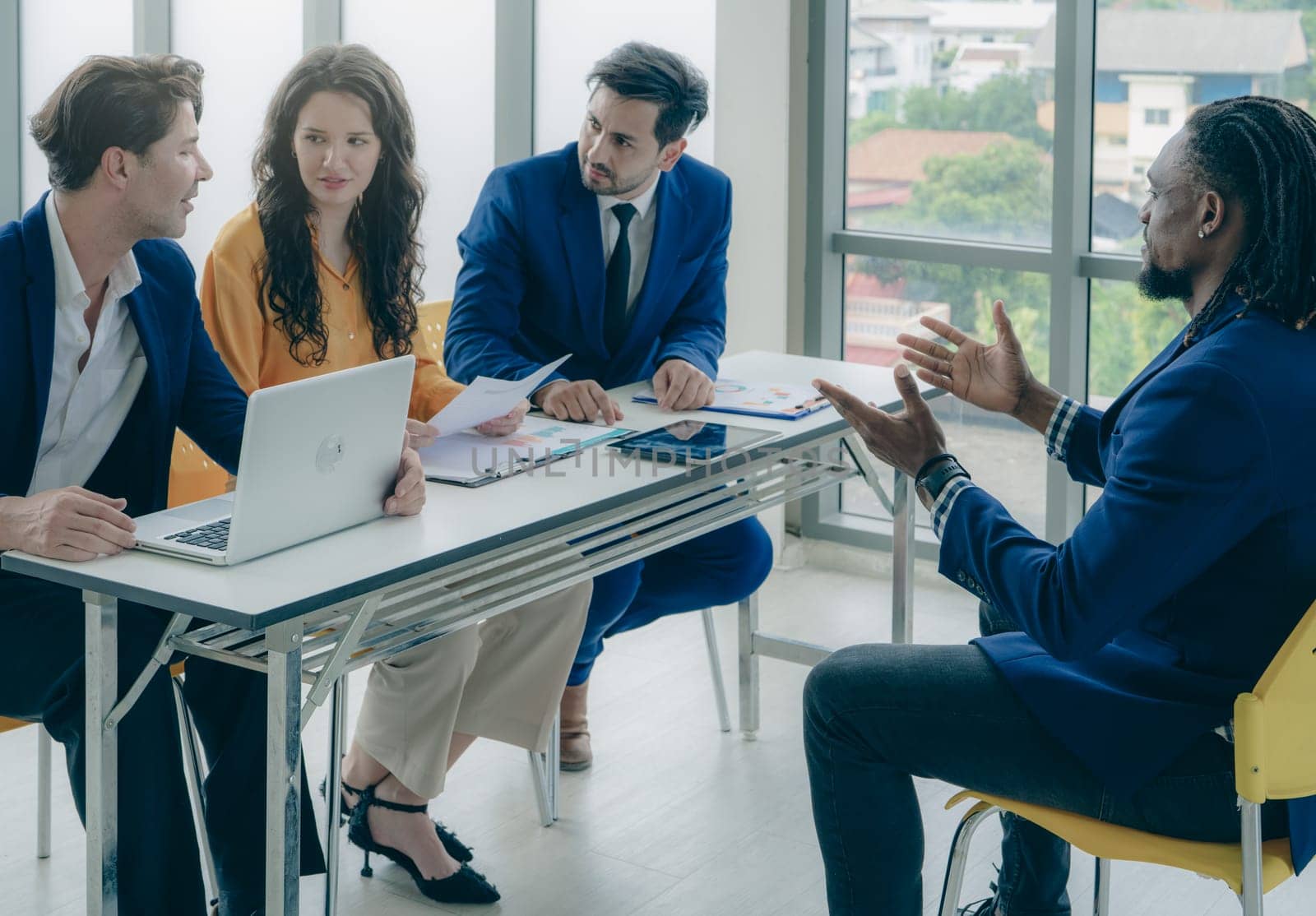 A group of people are sitting around a table in a business setting. The man in the blue suit is talking to the group. Scene is professional and serious