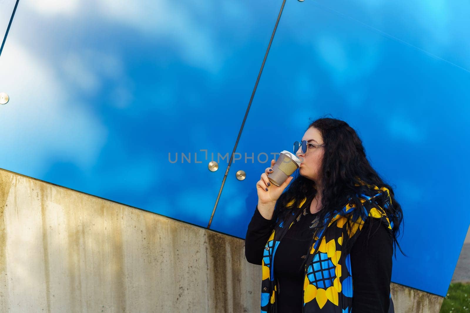 Beautiful young brunette woman with long curly hair, dressed in a black, blue and yellow waistcoat, drinks coffee from a paper cup on a background of blue wall.