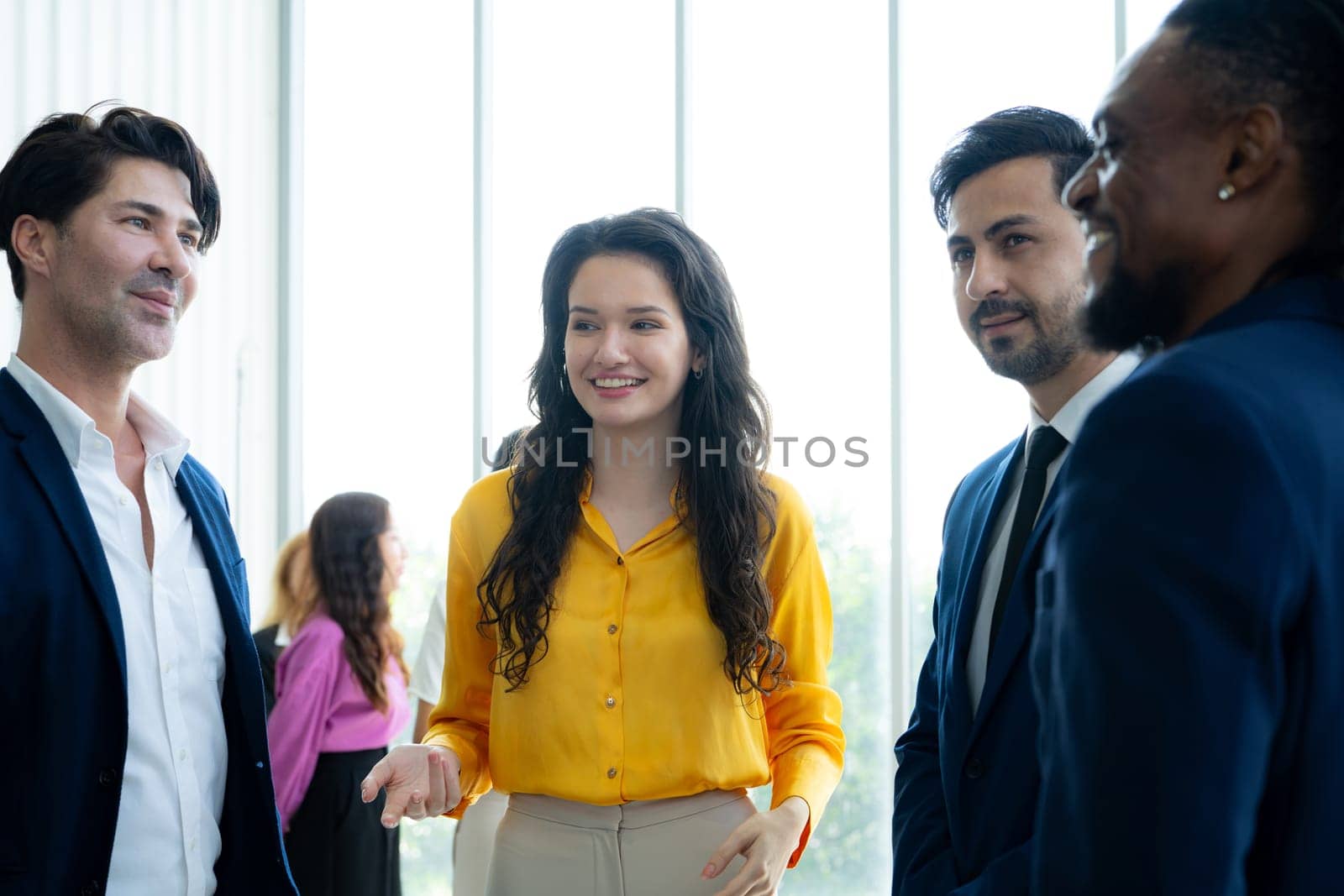 A group of people are standing together in a room, with one woman wearing a yellow shirt. The group appears to be friendly and engaged in conversation