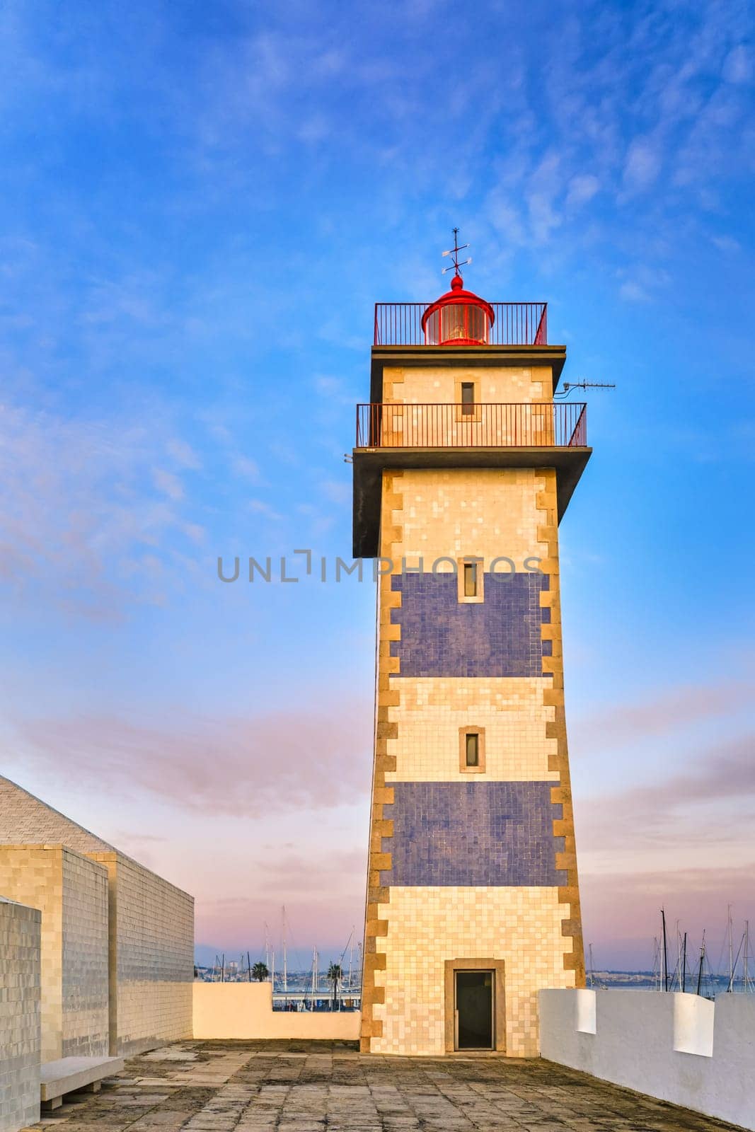 Upshot of Santa Marta lighthouse in Cascais, Portugal at sunset. Colorful sky, clouds, sunlight, low sun, calm ocean waters, local landmark, safety and navigation tower, rocky shore.
