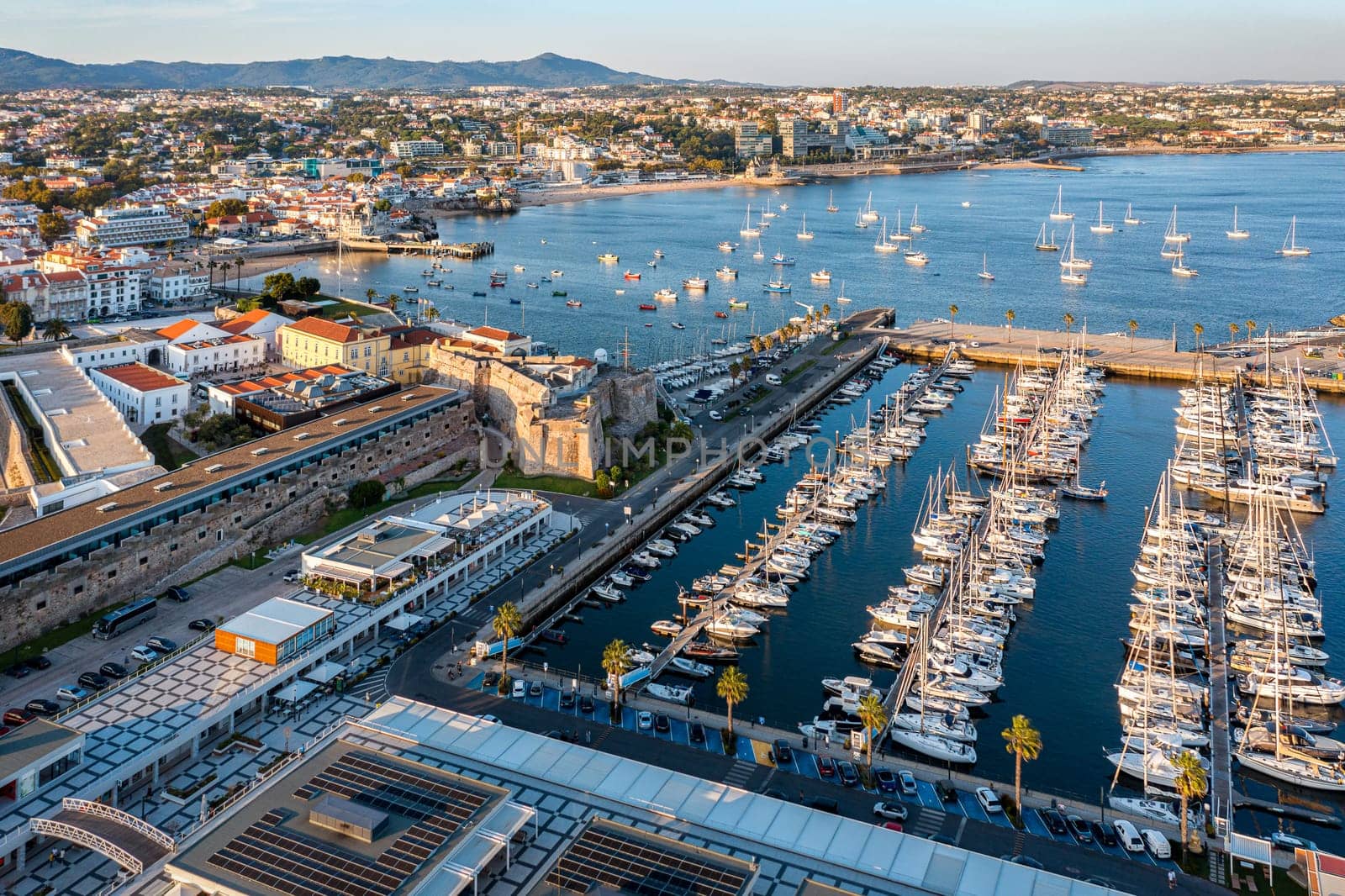 Drone view Cascais, coastal town, marina near Lisbon at sunny summer evening, drone point of view. Waters of the Atlantic Ocean, rocky coastline, sailboats, yachts, catamarans moored by jetties, low sun, luxury and leisure.