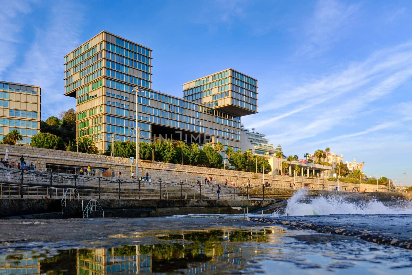 FEB 17, 2024 - Cascais, Portugal: Estoril Sol Residence, luxury block of apartments by waterfront, ocean pool and next to railway to Lisbon on sunny day, tidal waves splash on promenade. Modern architecture by Goncalo Byrne Arquitectos
