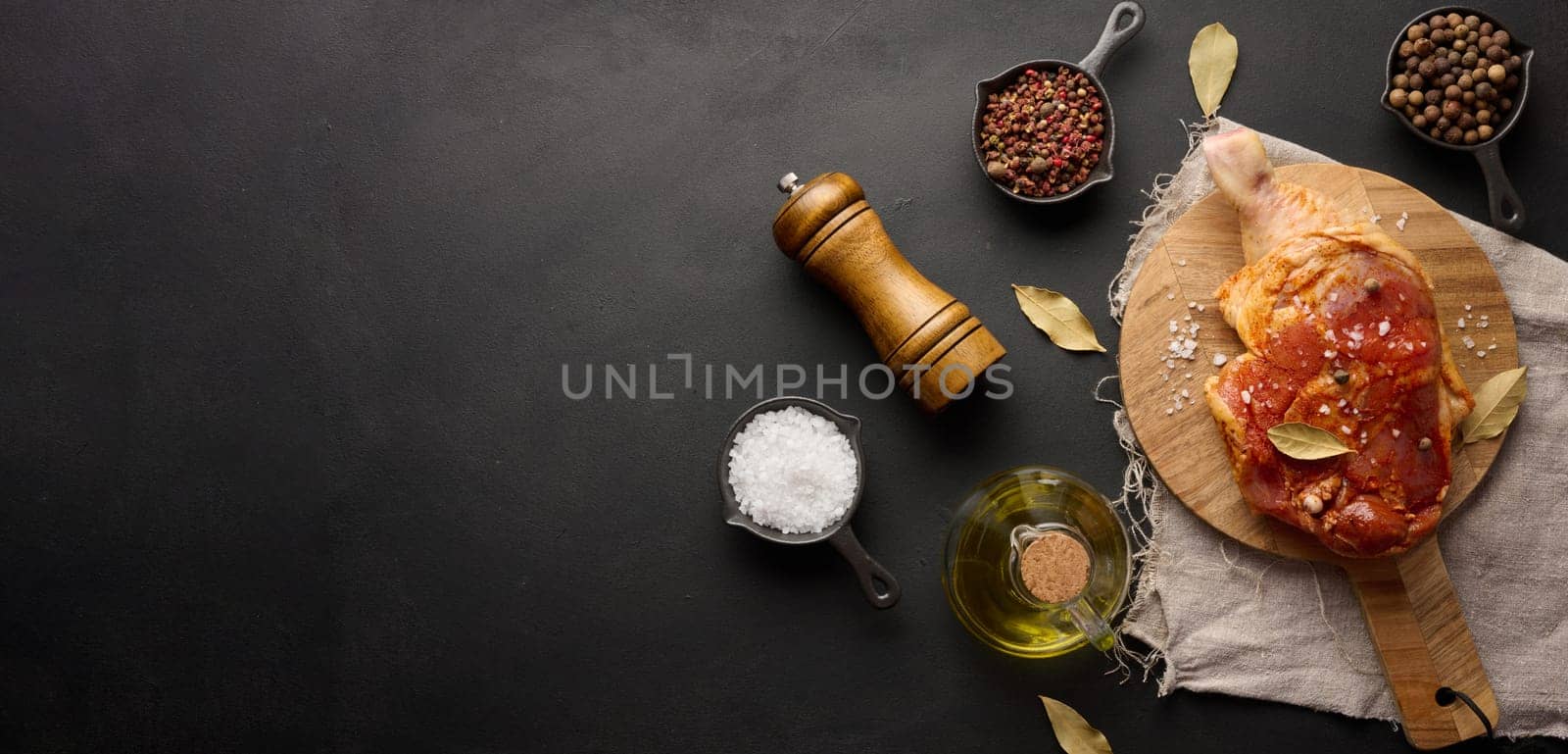 Raw chicken drumsticks seasoned on a wooden board, accompanied by salt and peppercorns, viewed from the top. Black table. Copy space