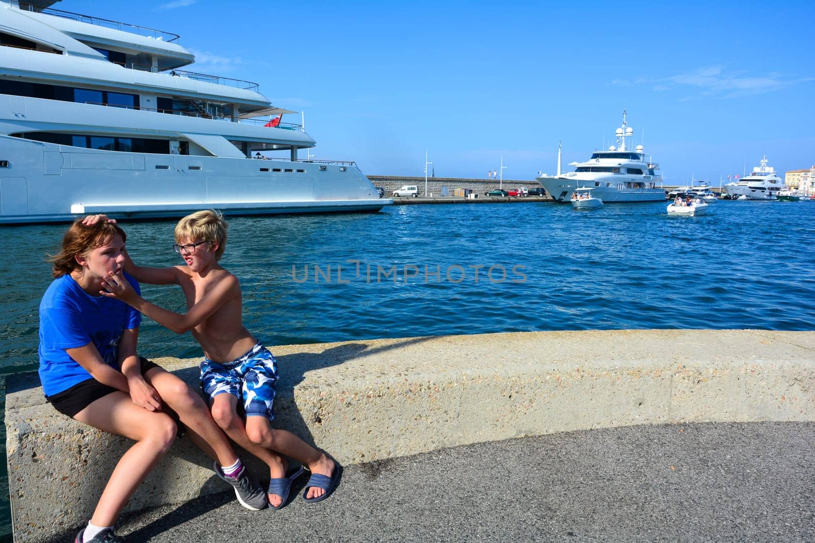 A girl and a boy sit on a wall at the harbor with funny facial expressions, big ships in the background