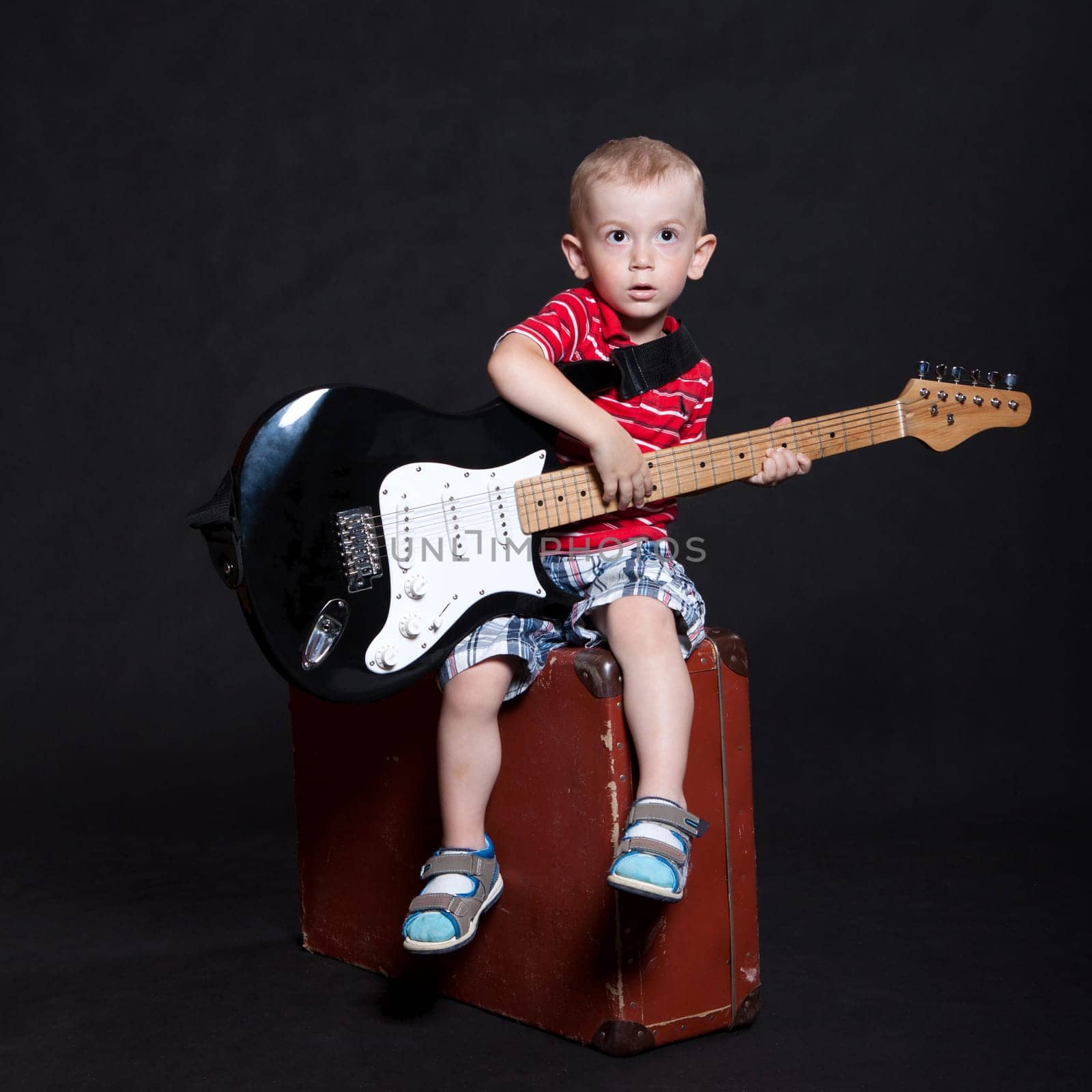 Small boy in striped shirt and checkered shorts sits on an old suitcase with an electric guitar