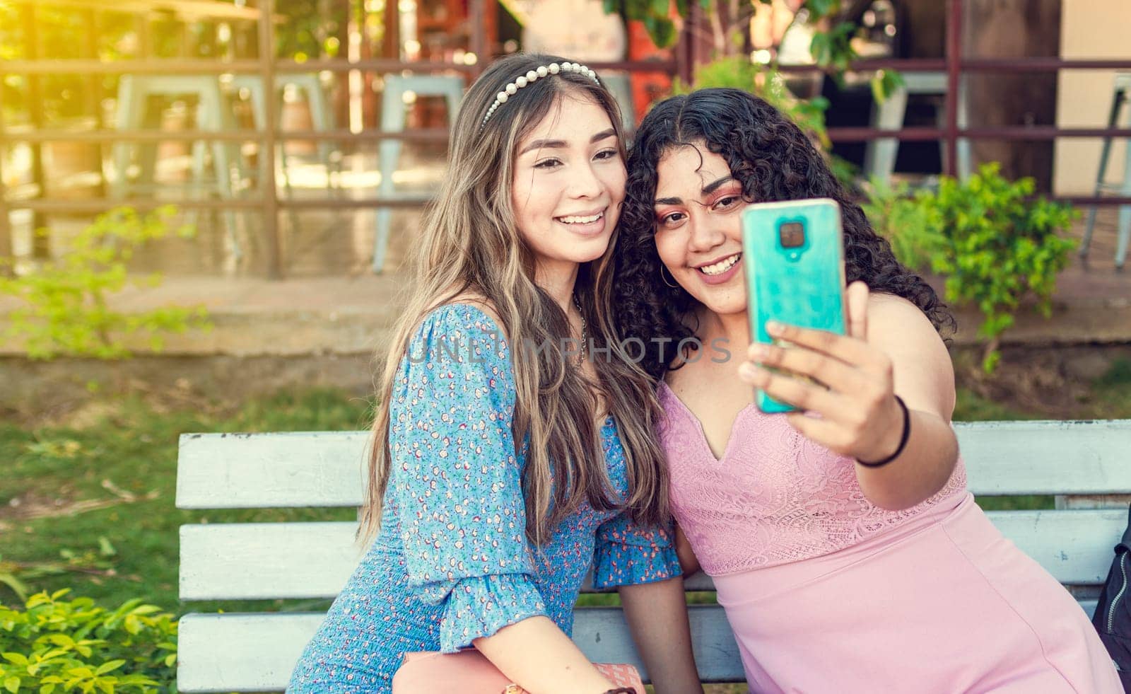 Two smiling teenage girls sitting on a bench taking a selfie, Two smiling teenage friends taking a selfie sitting on a bench at sunset