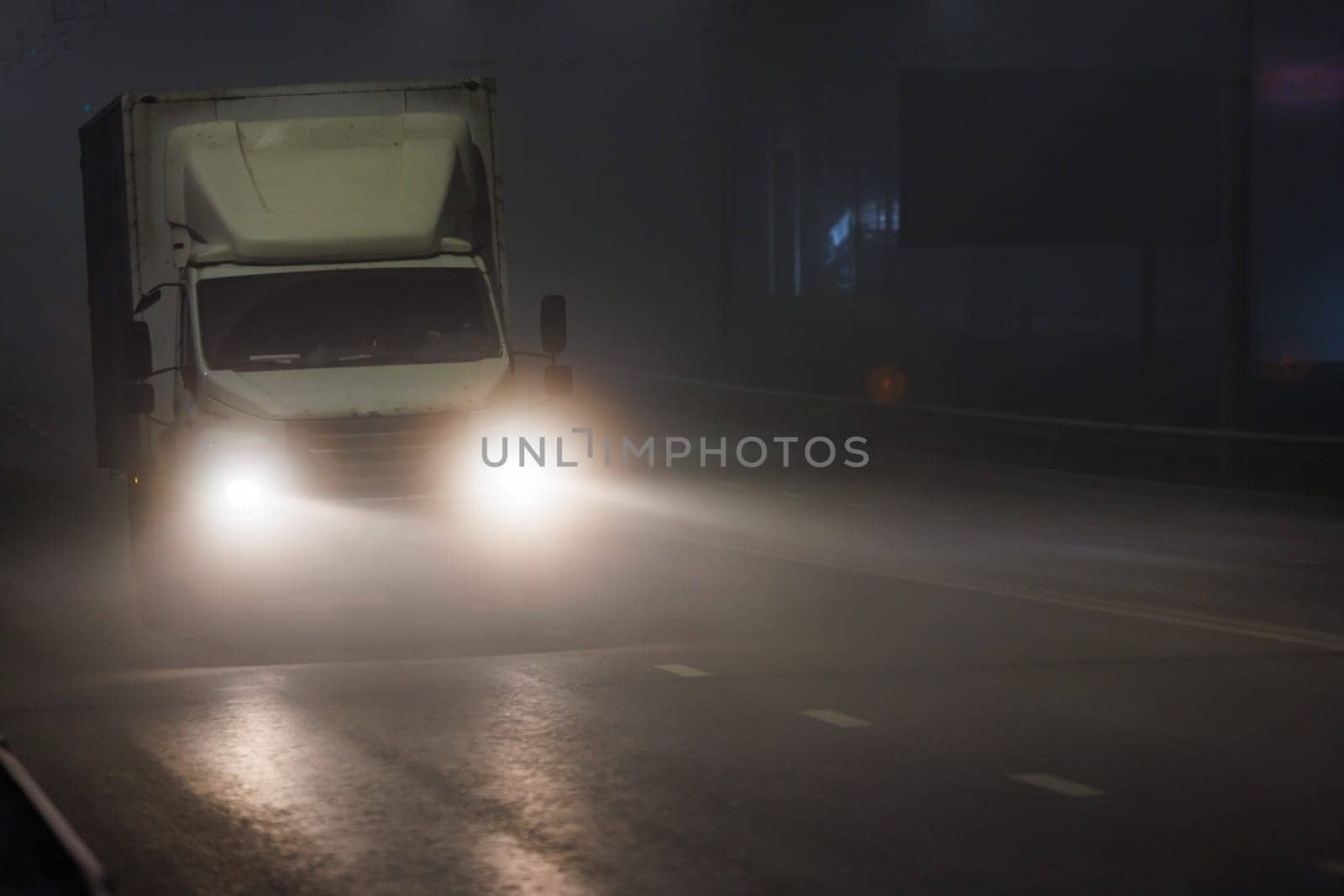 A vehicle with automotive lighting is rolling down the foggy highway at midnight. The headlamps illuminate the asphalt road surface as it travels through the dark night