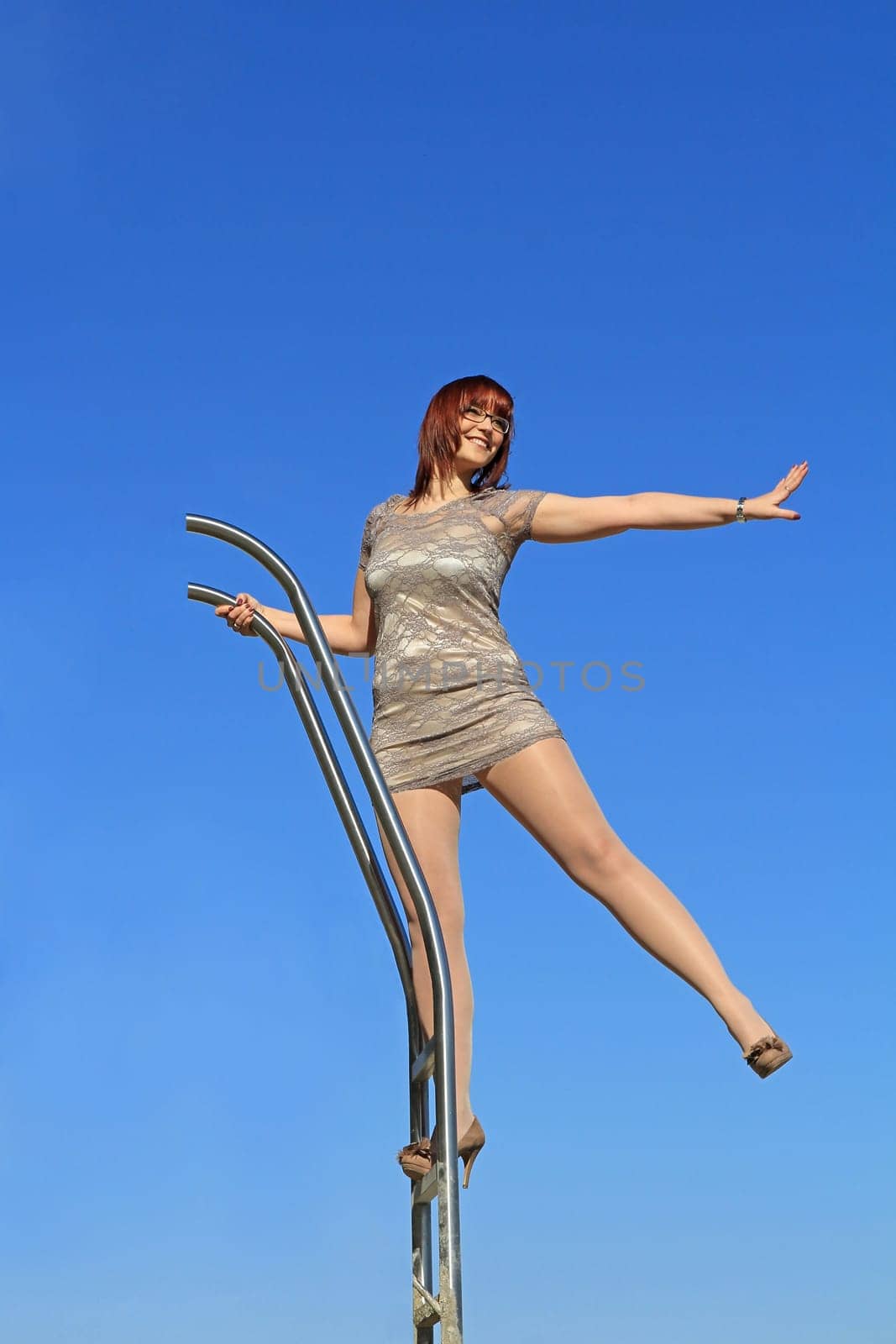 A young woman in a dress stands high up on a ladder and stretches out one arm and one leg