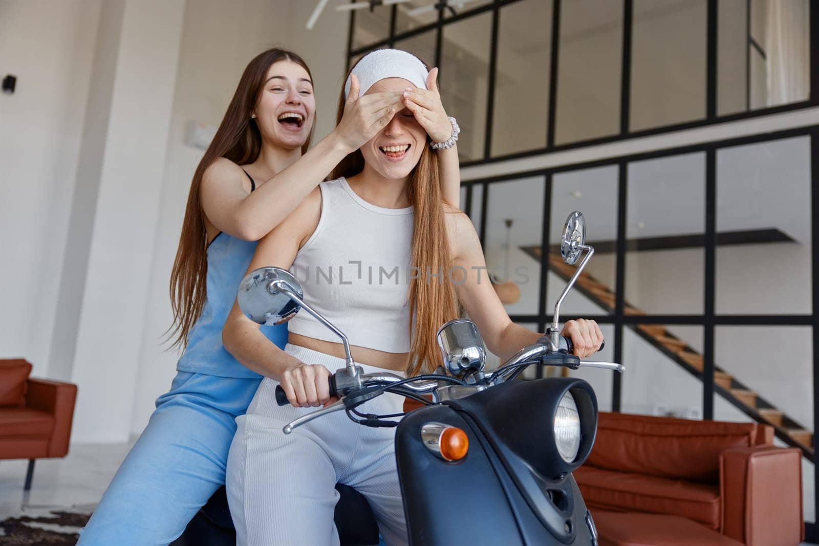 Two carefree girls having fun while sitting on motorbike over home interior. Youth culture and female beauty concept