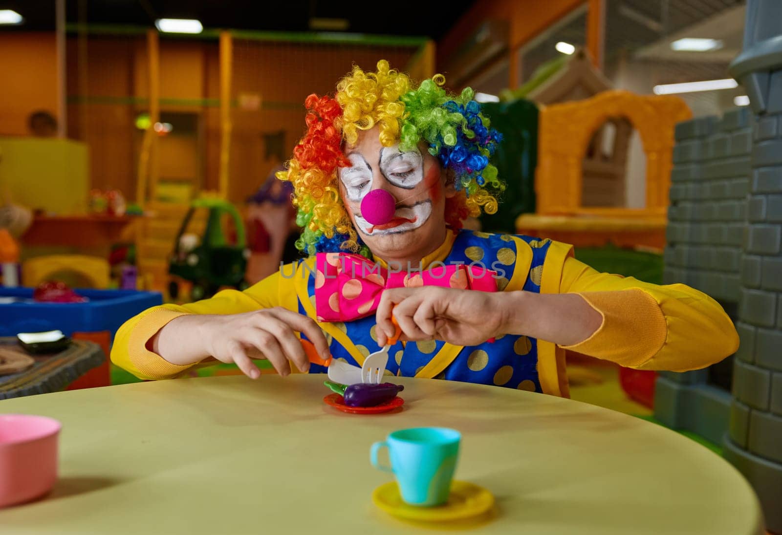 Funny clown wearing multi-colored wig and costume playing with toy tea set at kids playroom. Birthday party animation show concept